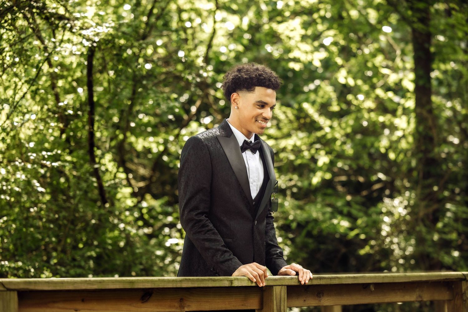 Someone in a black tuxedo and bowtie leans against a wooden railing in a lush green forest setting.