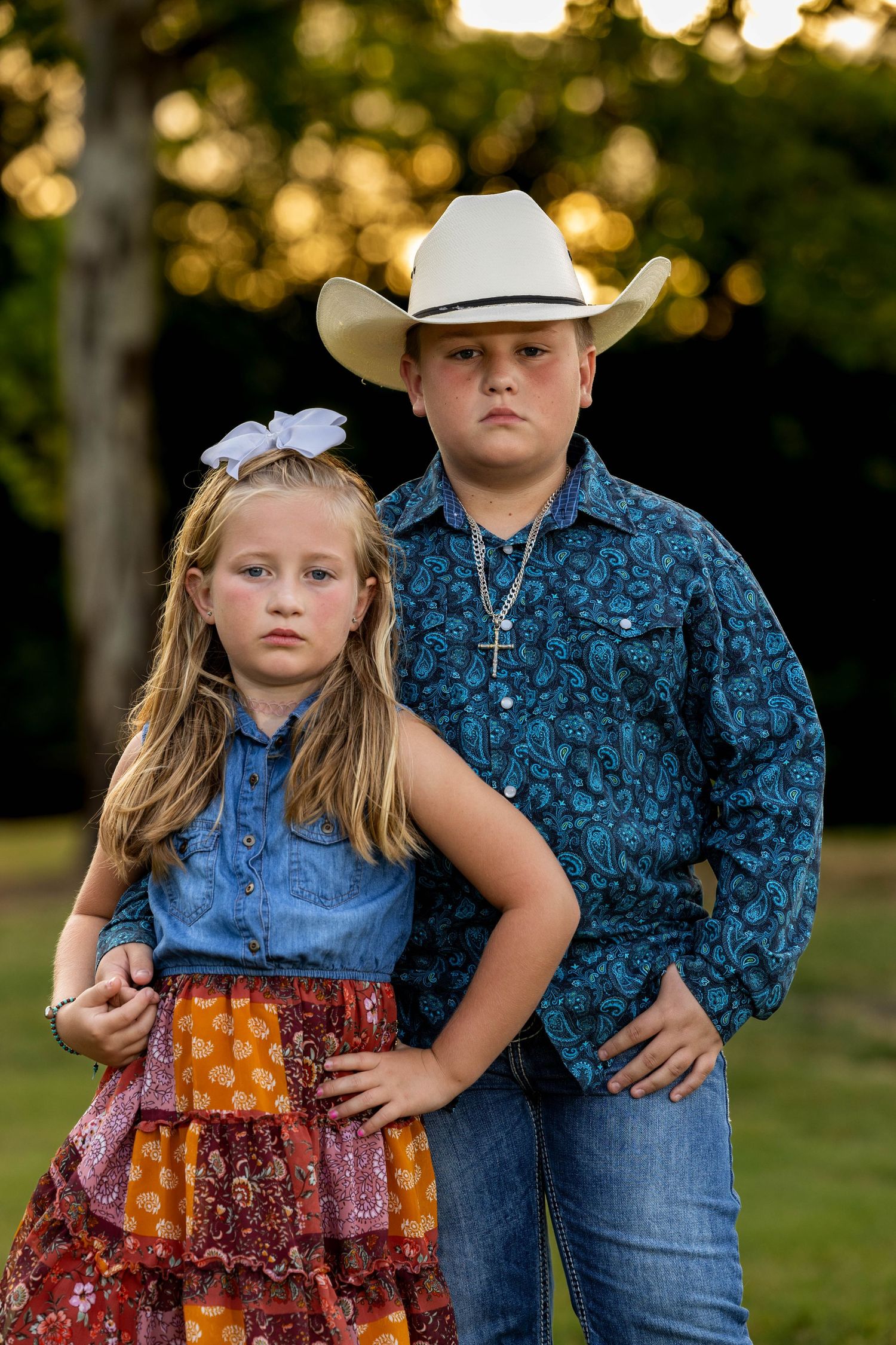 Two children in western attire pose confidently outdoors at golden hour.