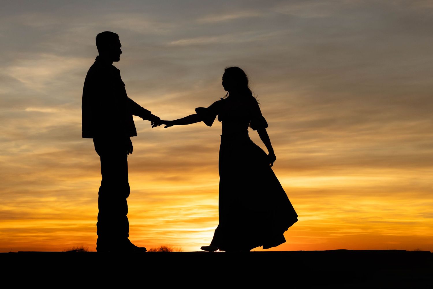 Two silhouettes hold hands against a dramatic orange sunset sky, creating a romantic scene on the horizon.