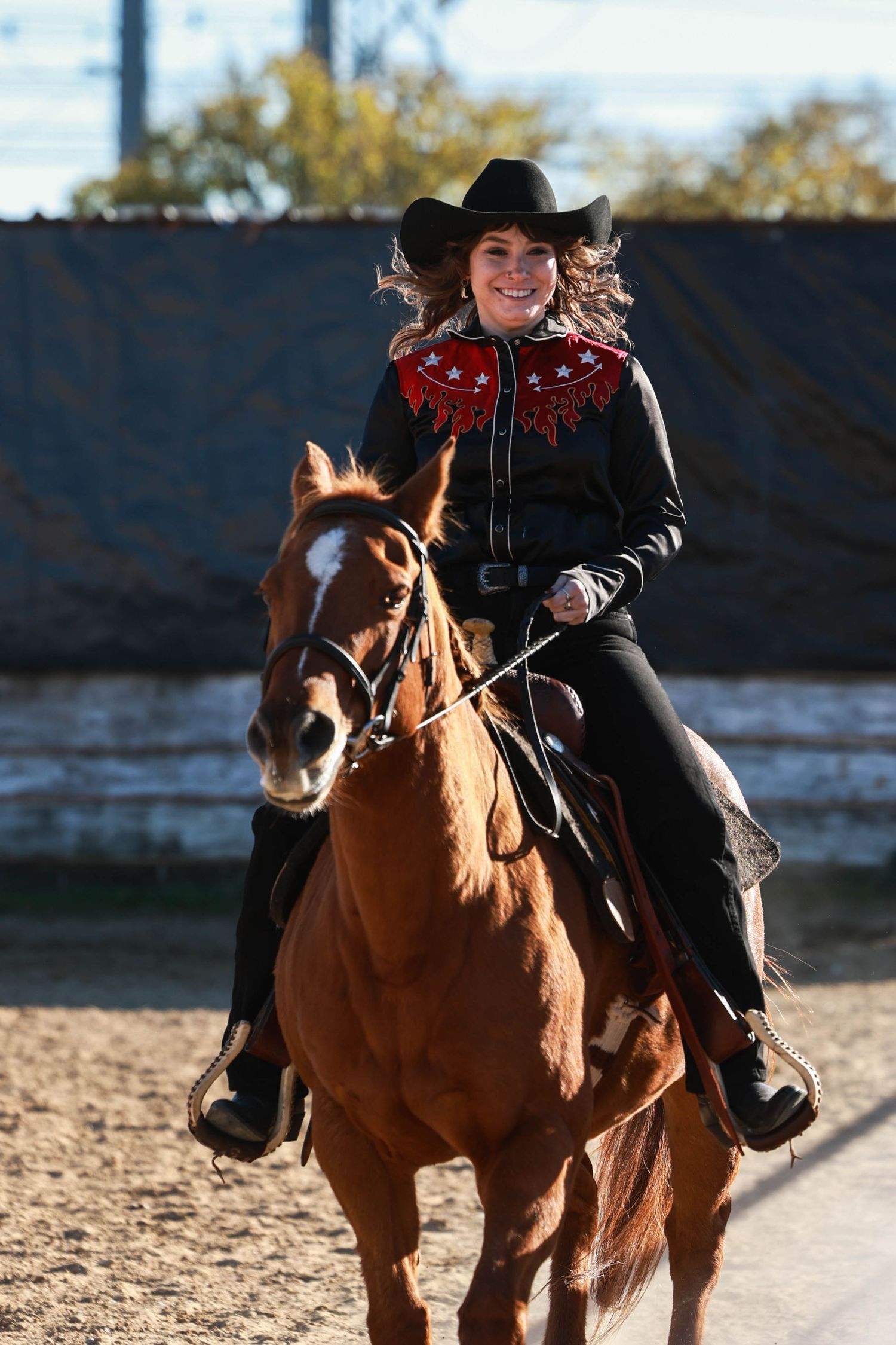 Rider in black western attire and red bandana rides a chestnut horse in an outdoor arena.