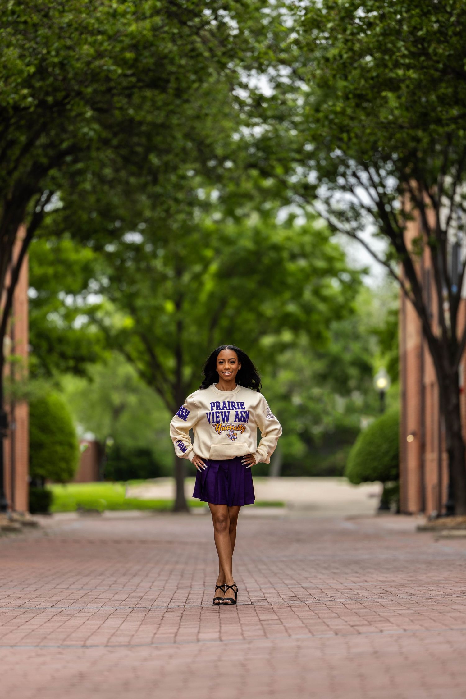 Person in university sweatshirt and skirt walks down tree-lined brick pathway.