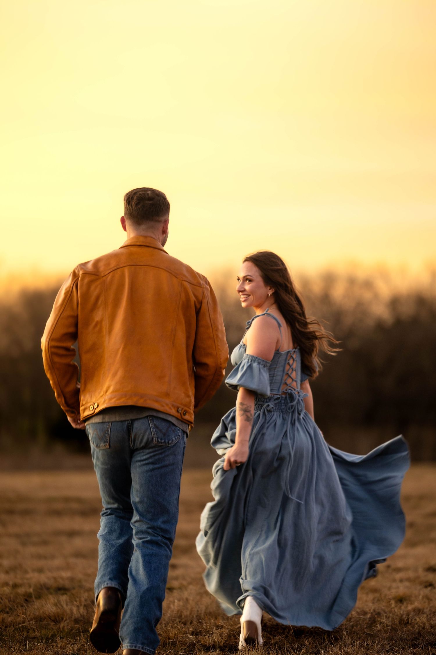 A couple walks through a field at sunset with her blue dress flowing in the wind.