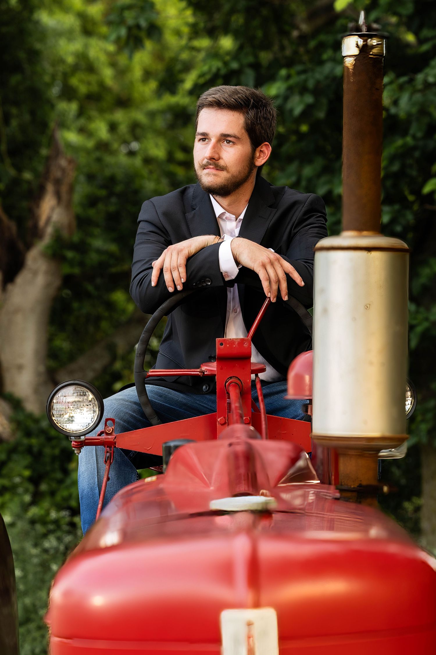 A suited person sits on a vintage red tractor in an outdoor setting with lush greenery.