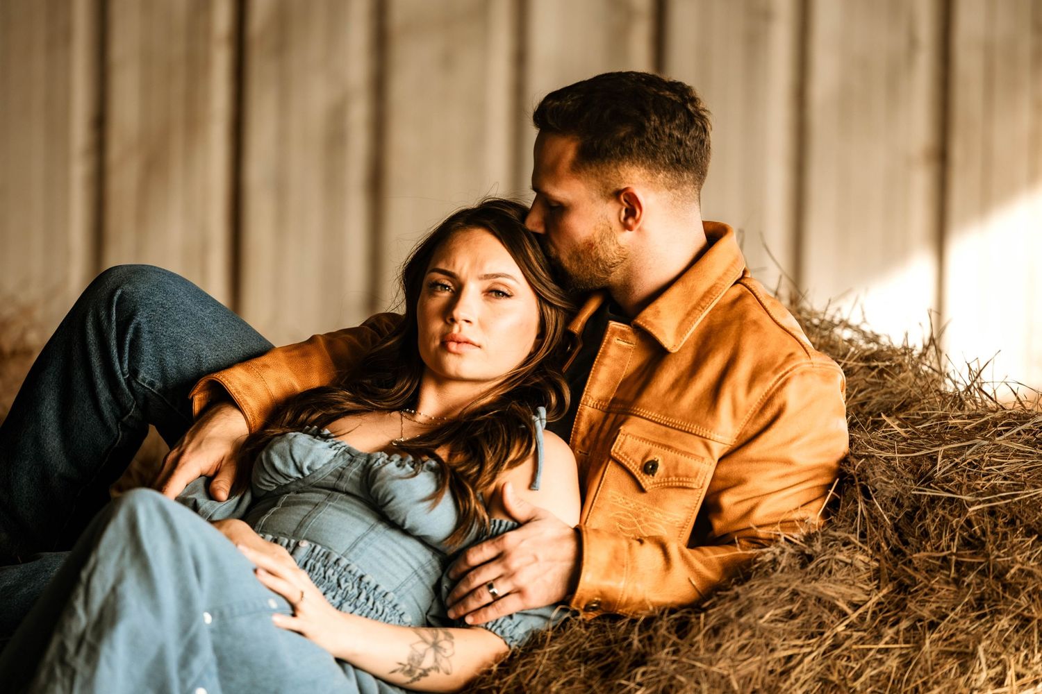 A romantic couple lounges on hay bales wearing denim and brown jacket.