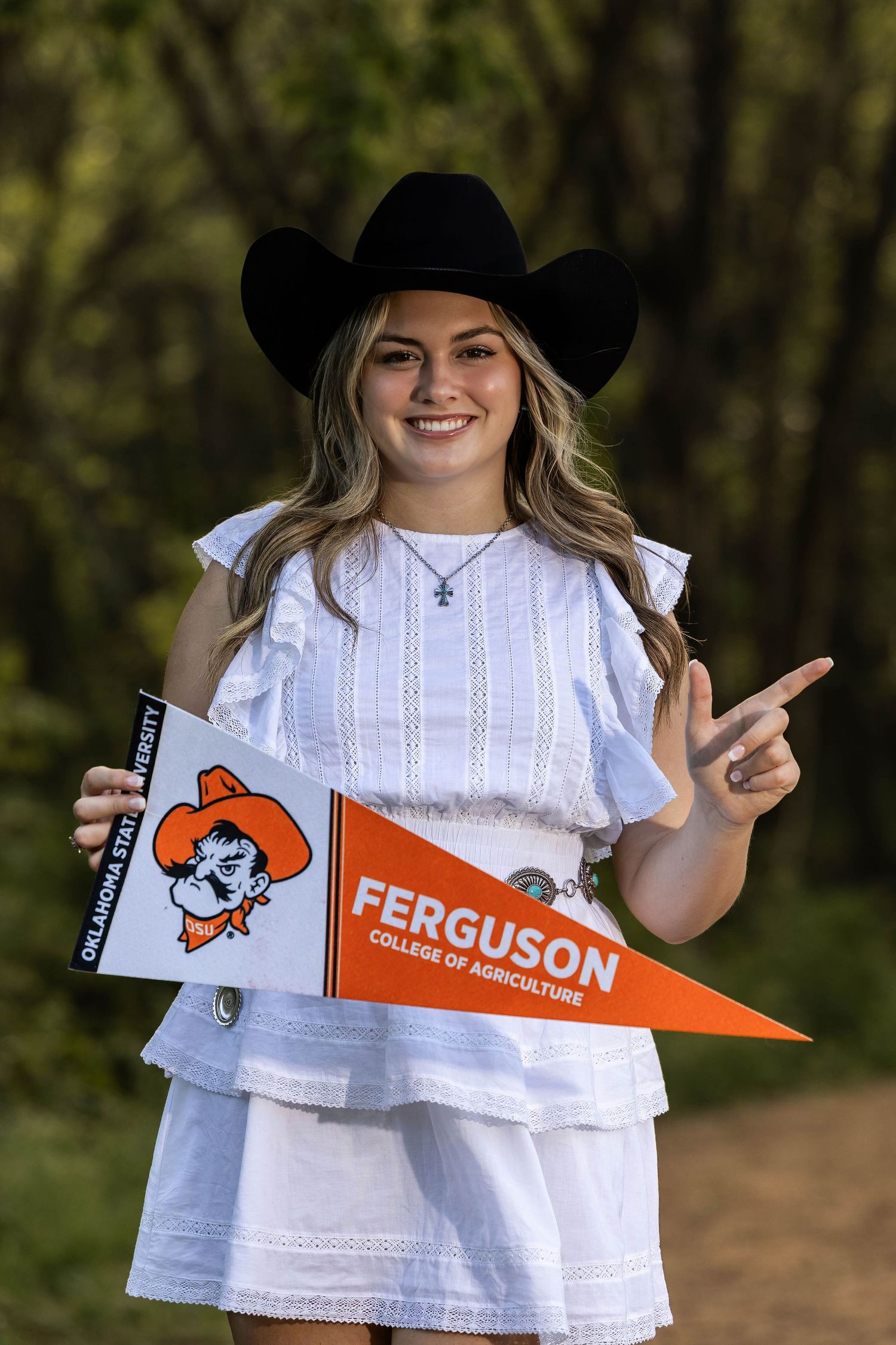 Person in white dress and cowboy hat holds Ferguson college pennant outdoors.