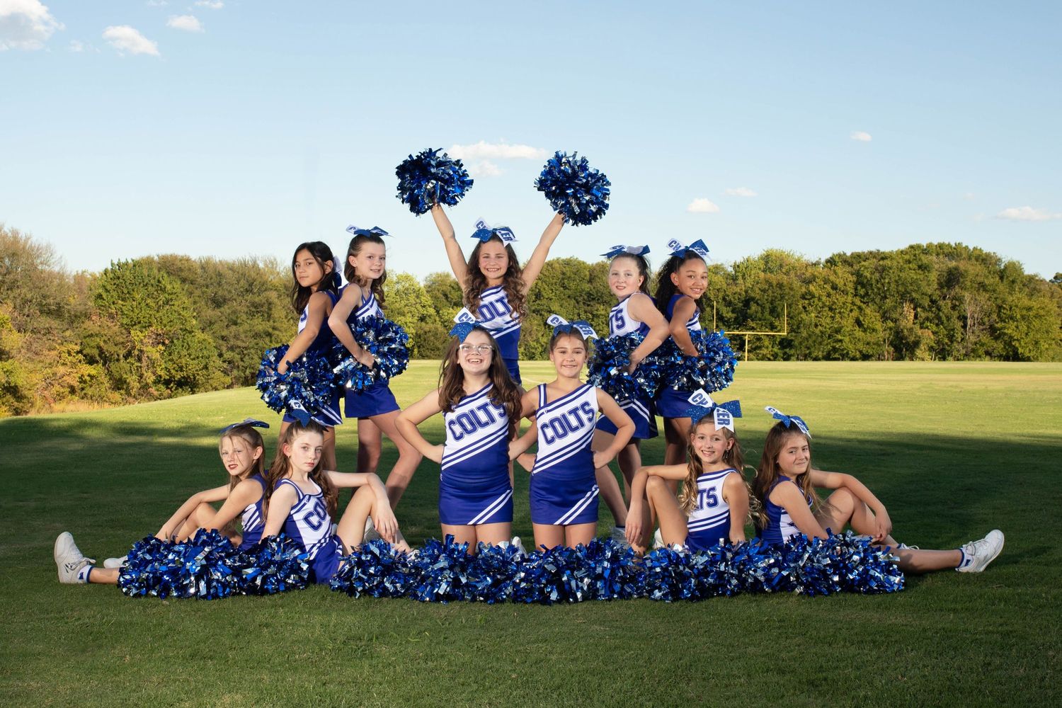 Cheerleading squad in blue and white uniforms poses together on grass field with pom poms and matching accessories.