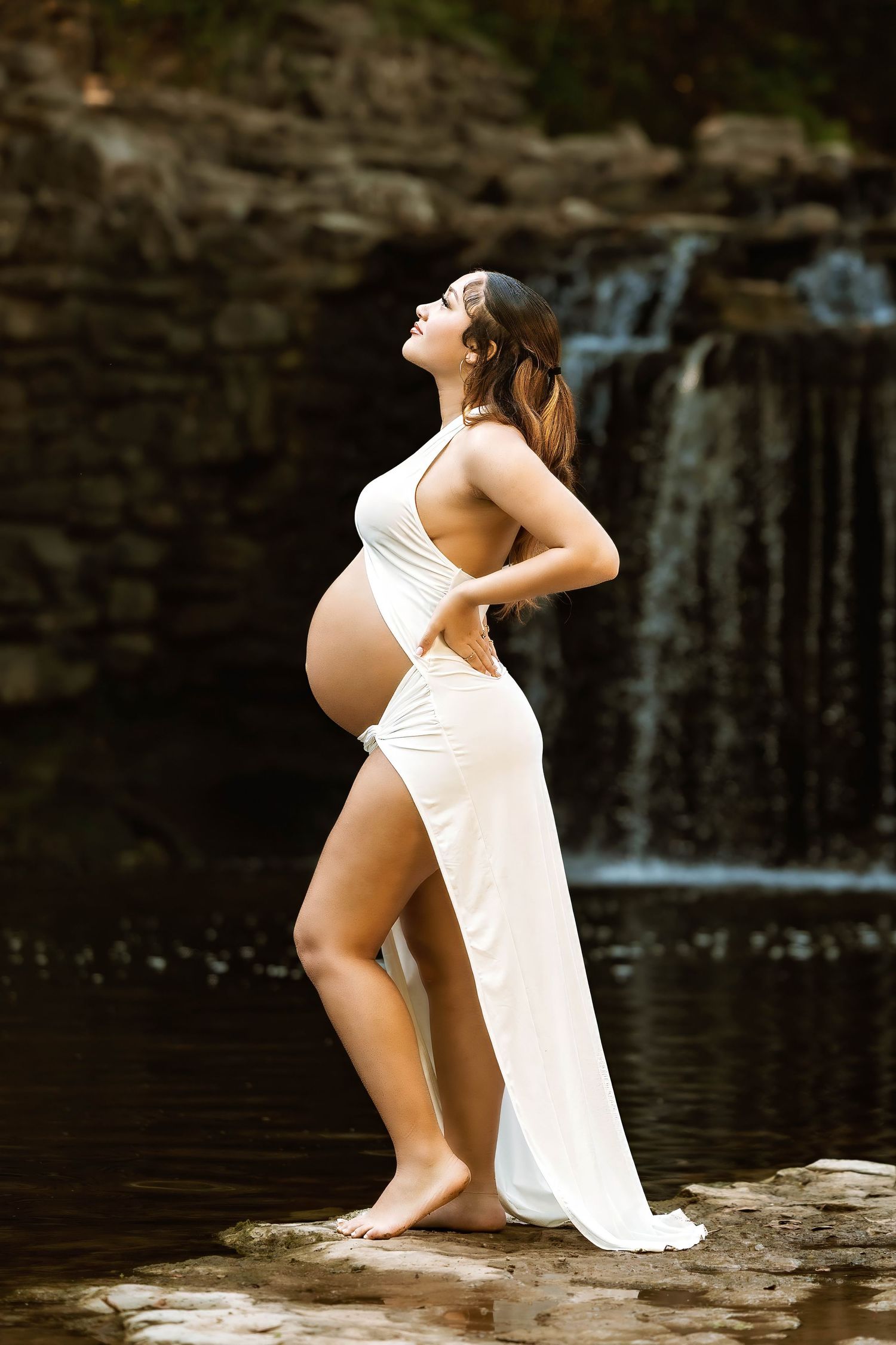 A pregnant woman in a white dress poses gracefully by a waterfall, arching her back in a serene maternity photoshoot.
