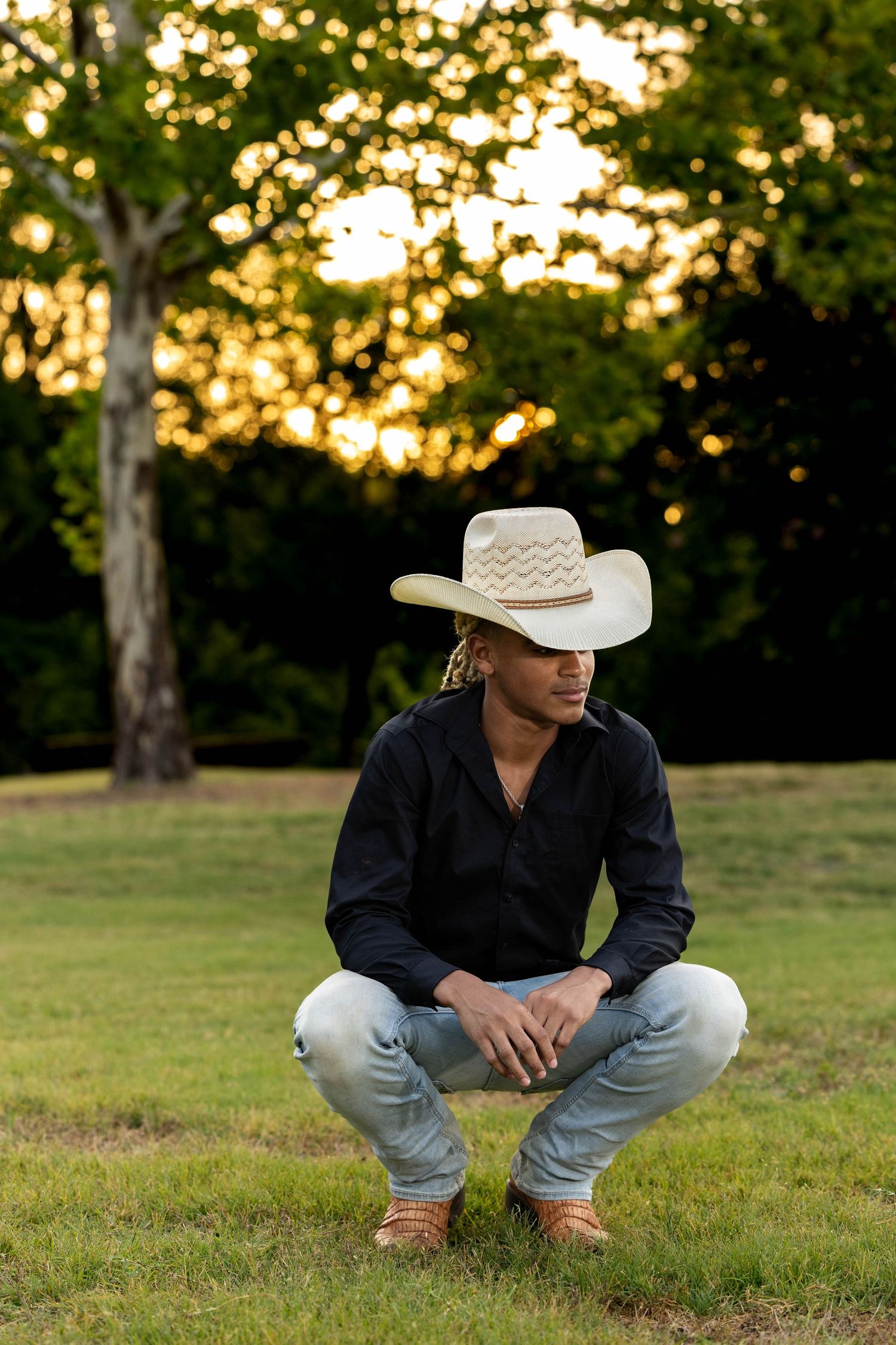 Person in cowboy hat and jeans crouches on grass during golden hour sunset.