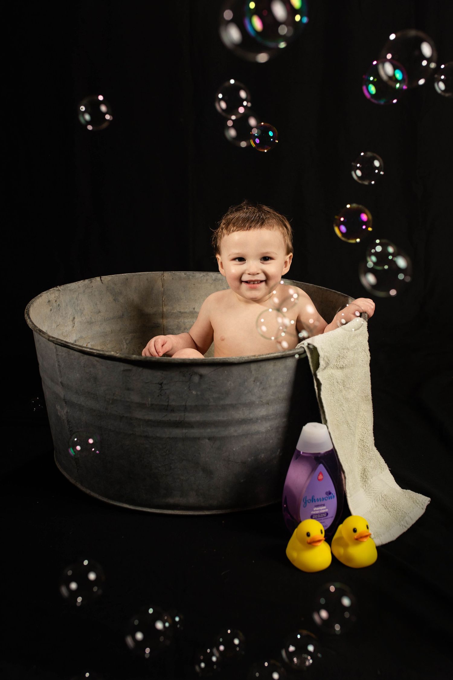 Baby sitting in vintage metal tub with rubber ducks and bubbles against black background.
