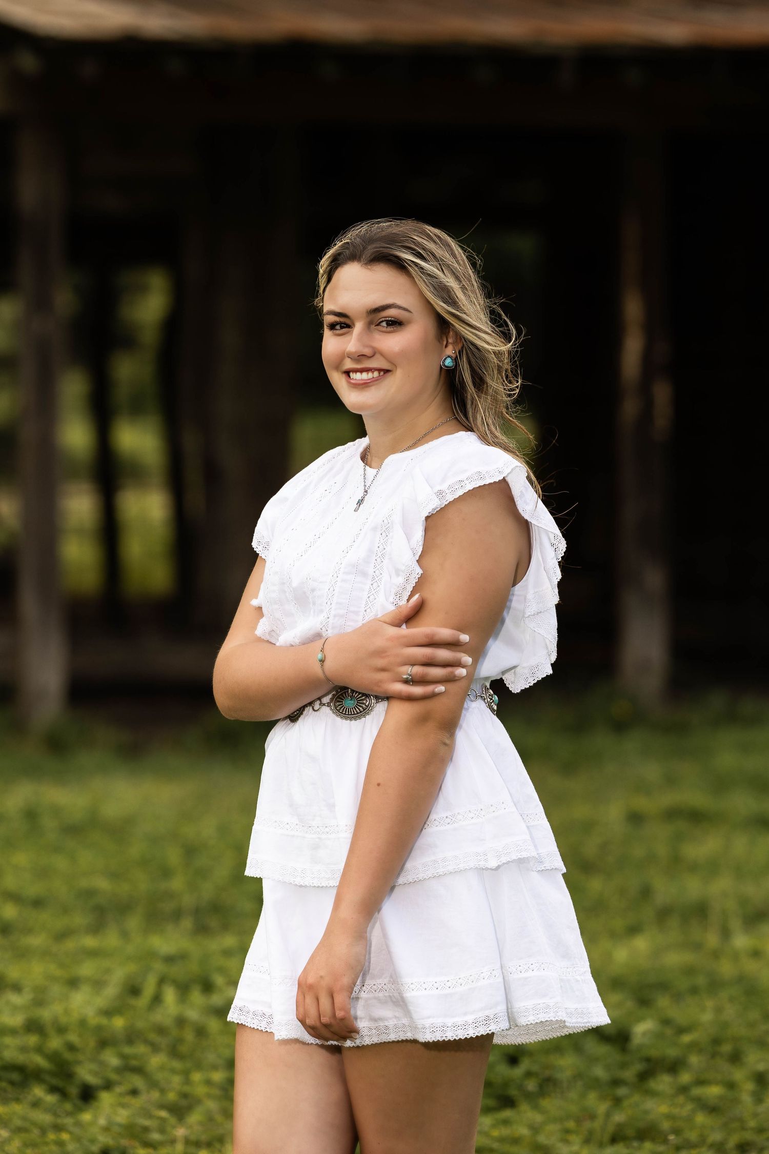 Person in white ruffle dress poses outdoors near rustic wooden structure.