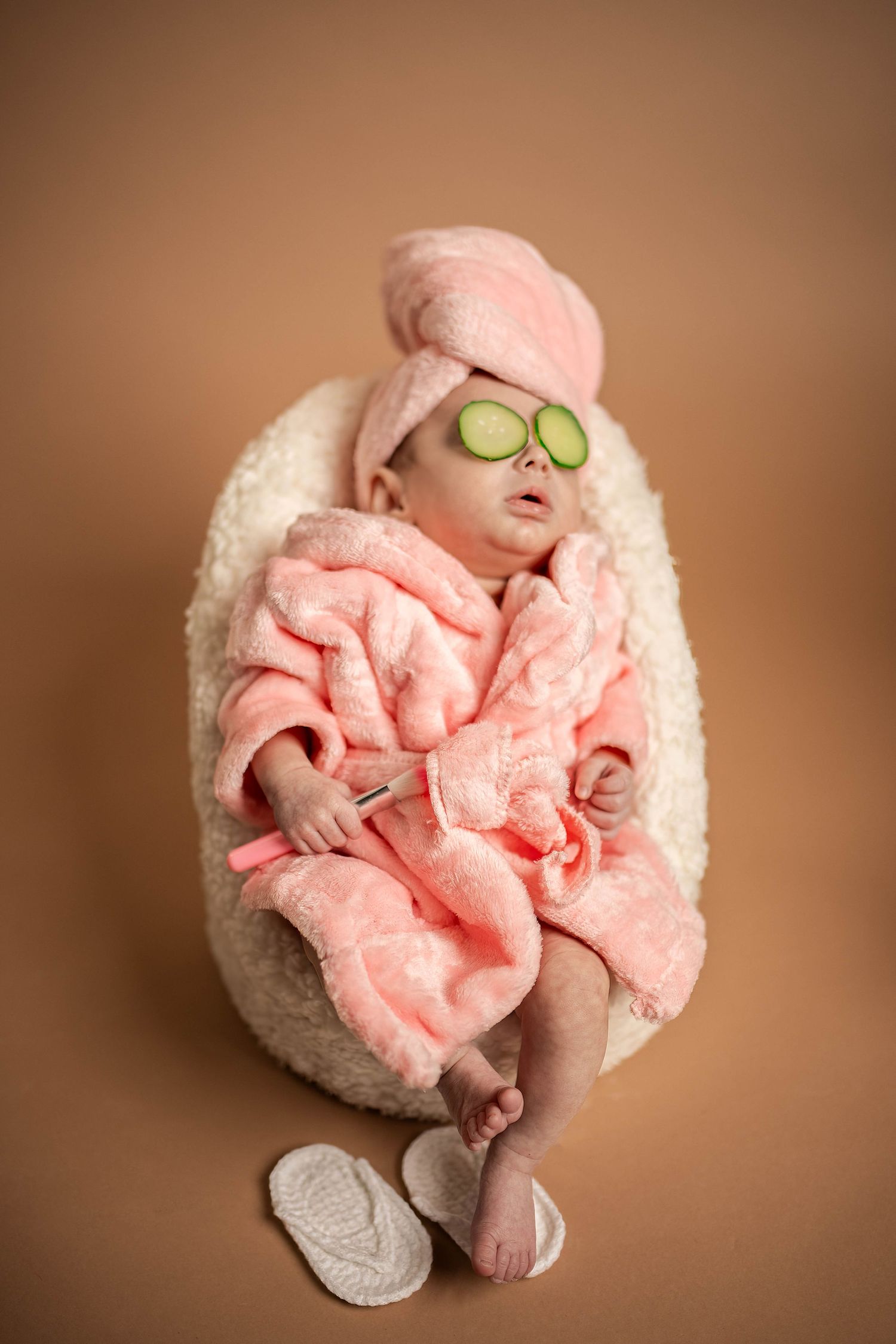 Newborn dressed in pink robe with cucumber slices on eyes in spa-themed photo shoot.