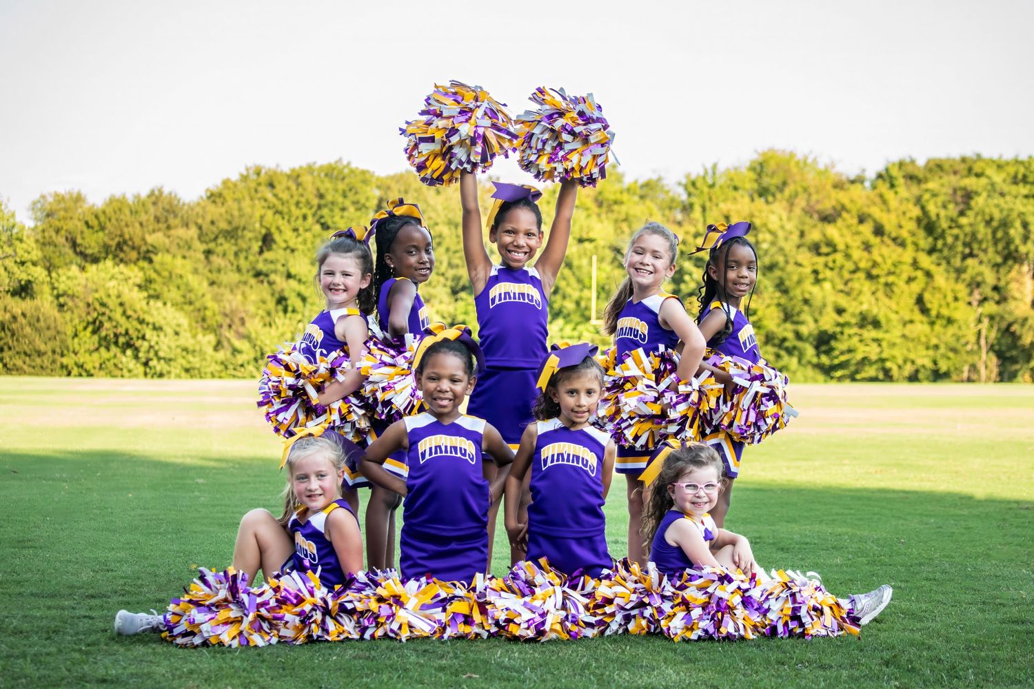 Cheerleaders in purple uniforms form a pyramidal formation on a grassy field with pom poms and smiles.