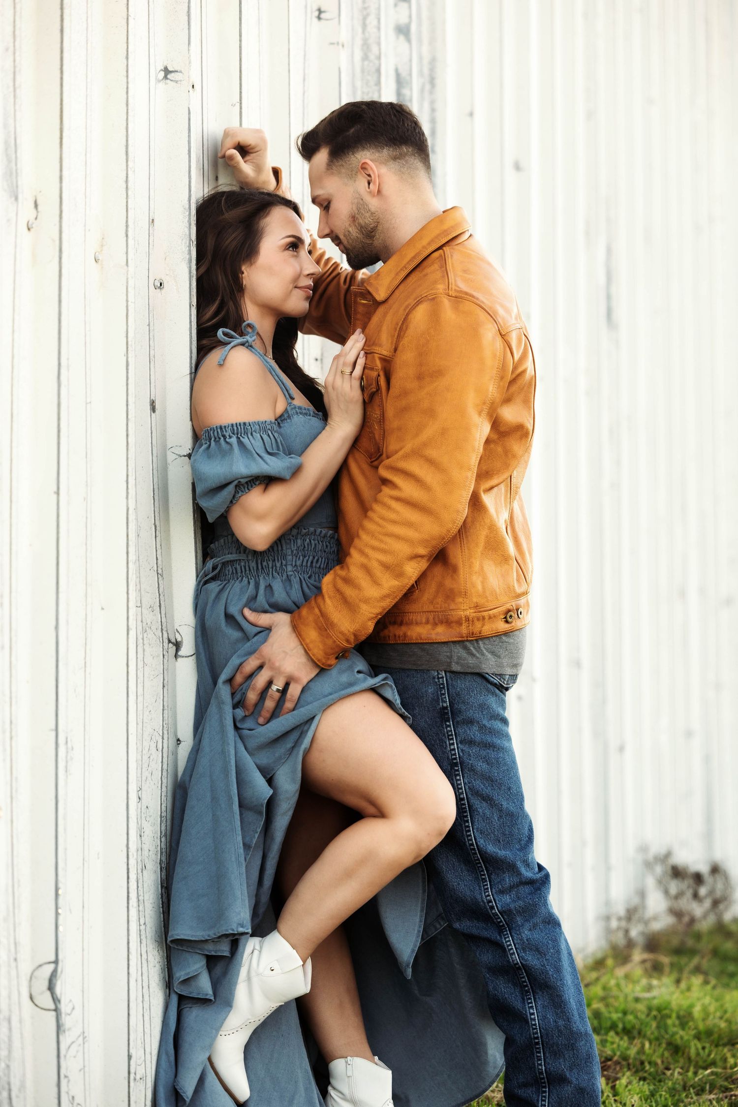 A romantic couple embraces against a white wall, she wears a blue dress and he wears an orange jacket.