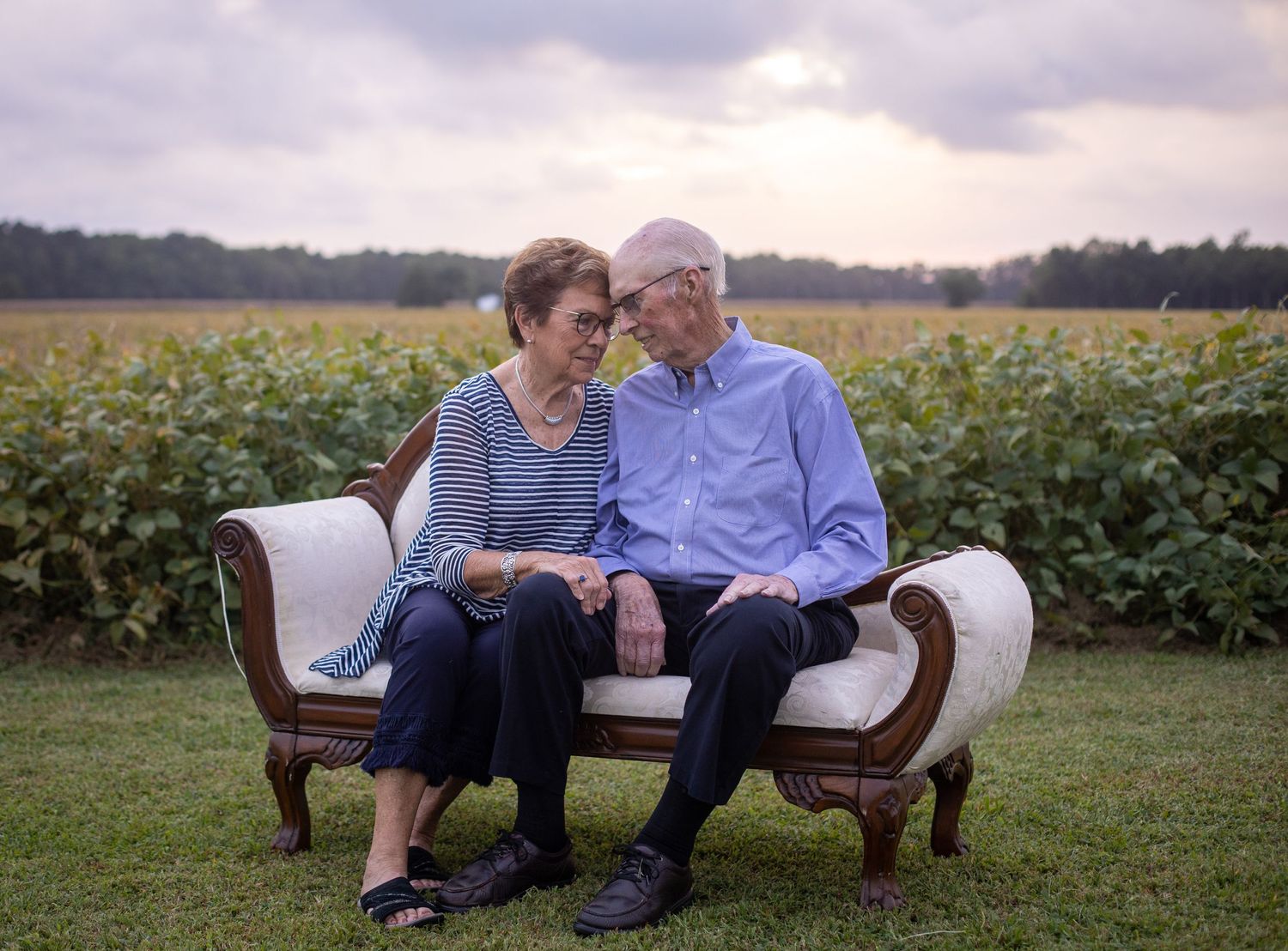 an elderly couple in their 80s touching foreheads with a field of soybeans in the background as the sun sets on a cloudy evening