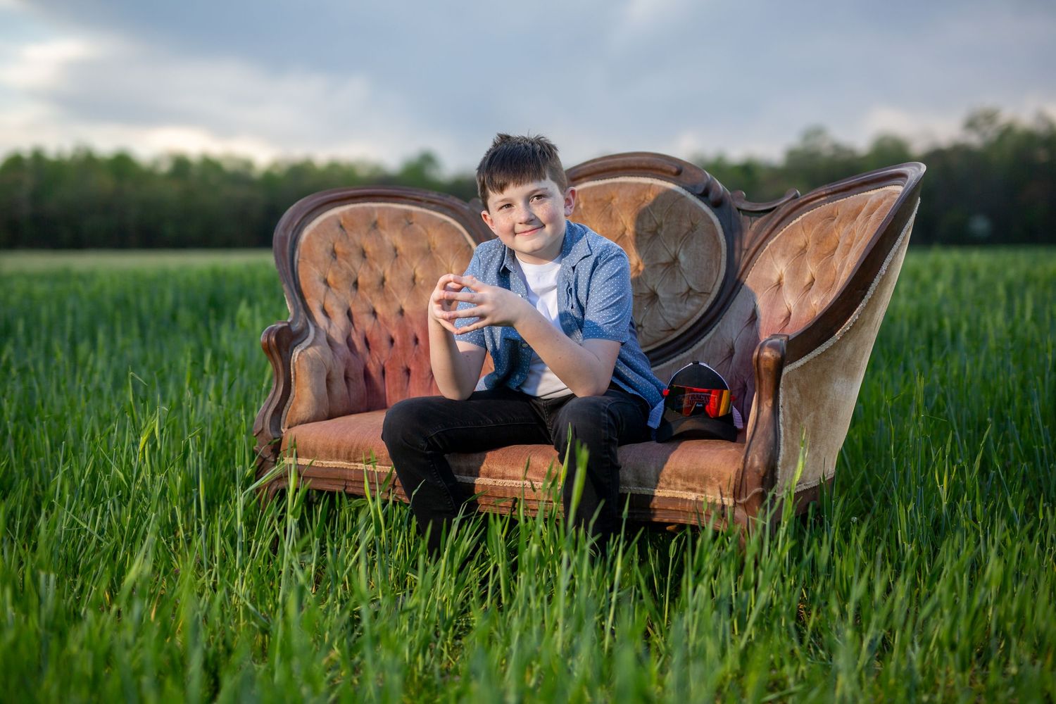 ten year old boy sitting on an antique couch in a field of tall green grass as the sun sets behind him photography by Ashley chance