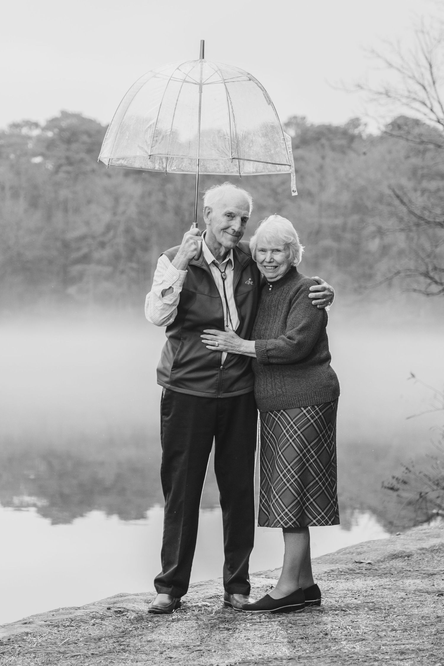 an elderly couple in their 80s by a lake with fog around them holding an umbrella and embracing photography by Ashley chance trap pond