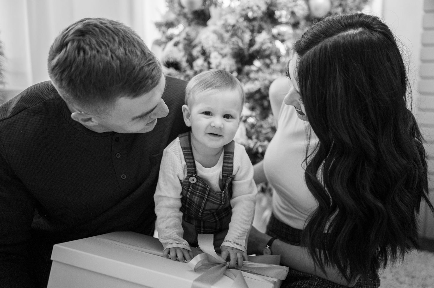 A baby in overalls stands between parents in a black and white holiday photo with Christmas tree.