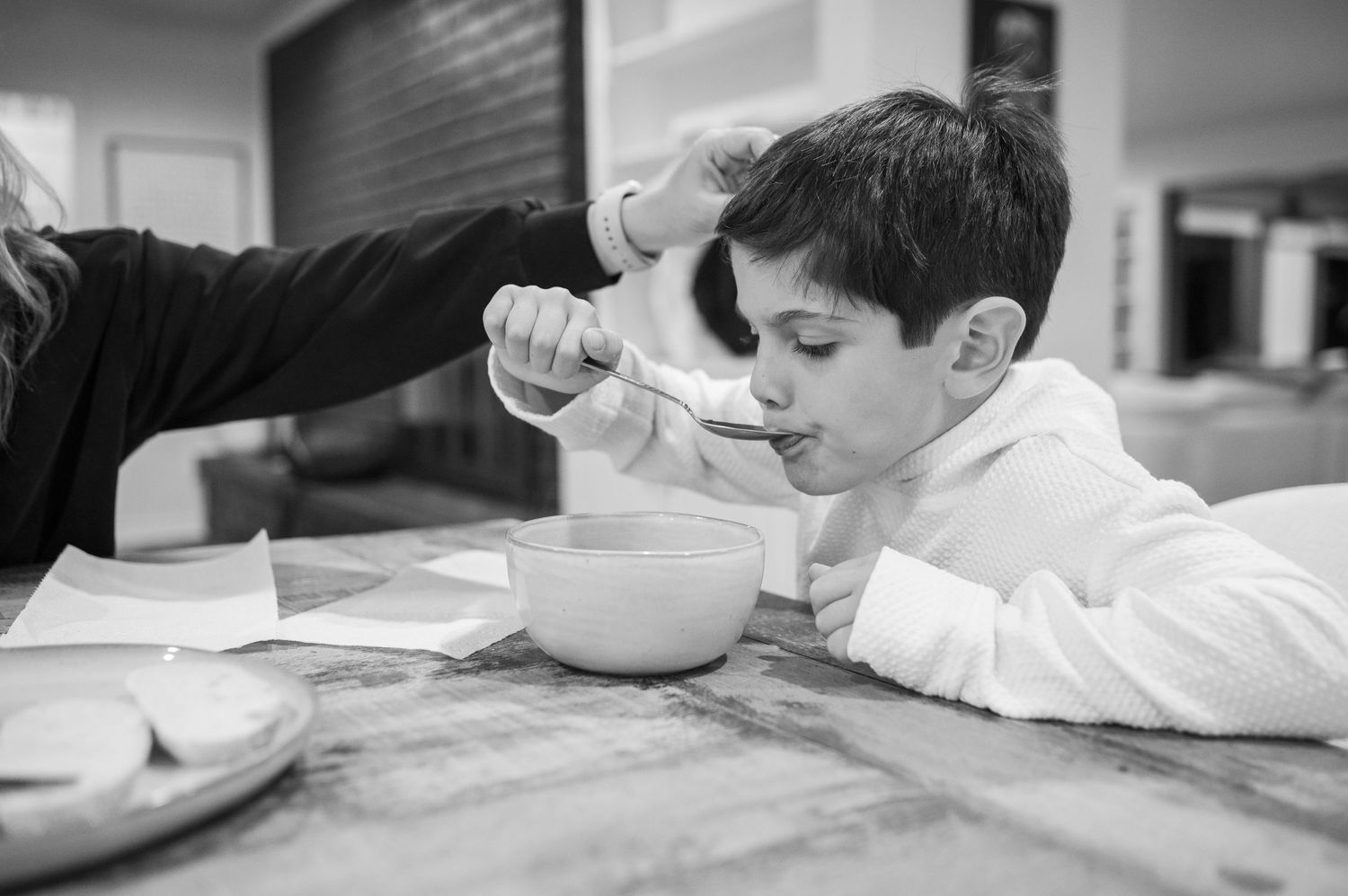 A young child in white eating from a bowl in the kitchen.