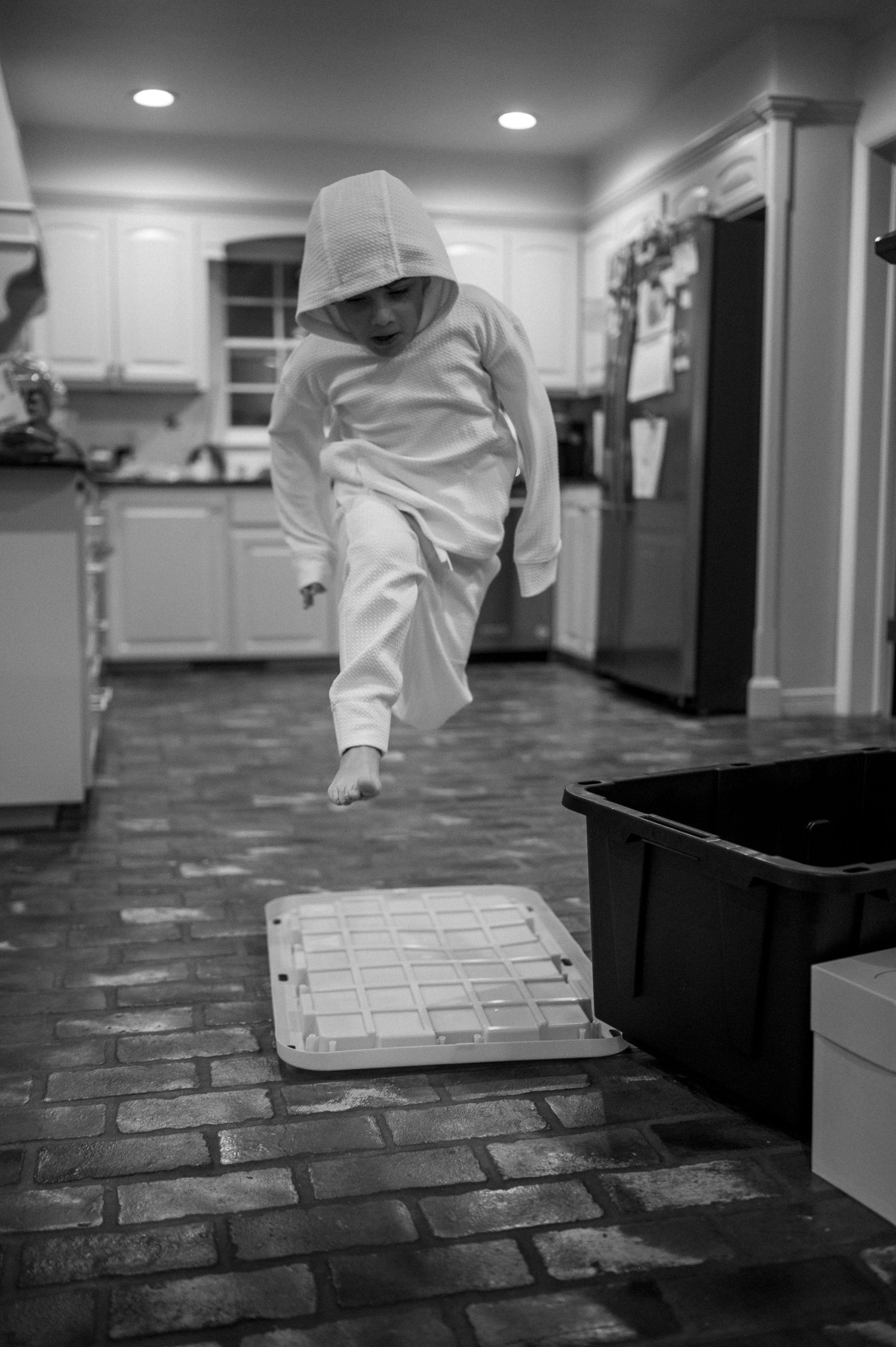 A person in white clothing performs a playful jump in a modern kitchen with brick-style flooring.
