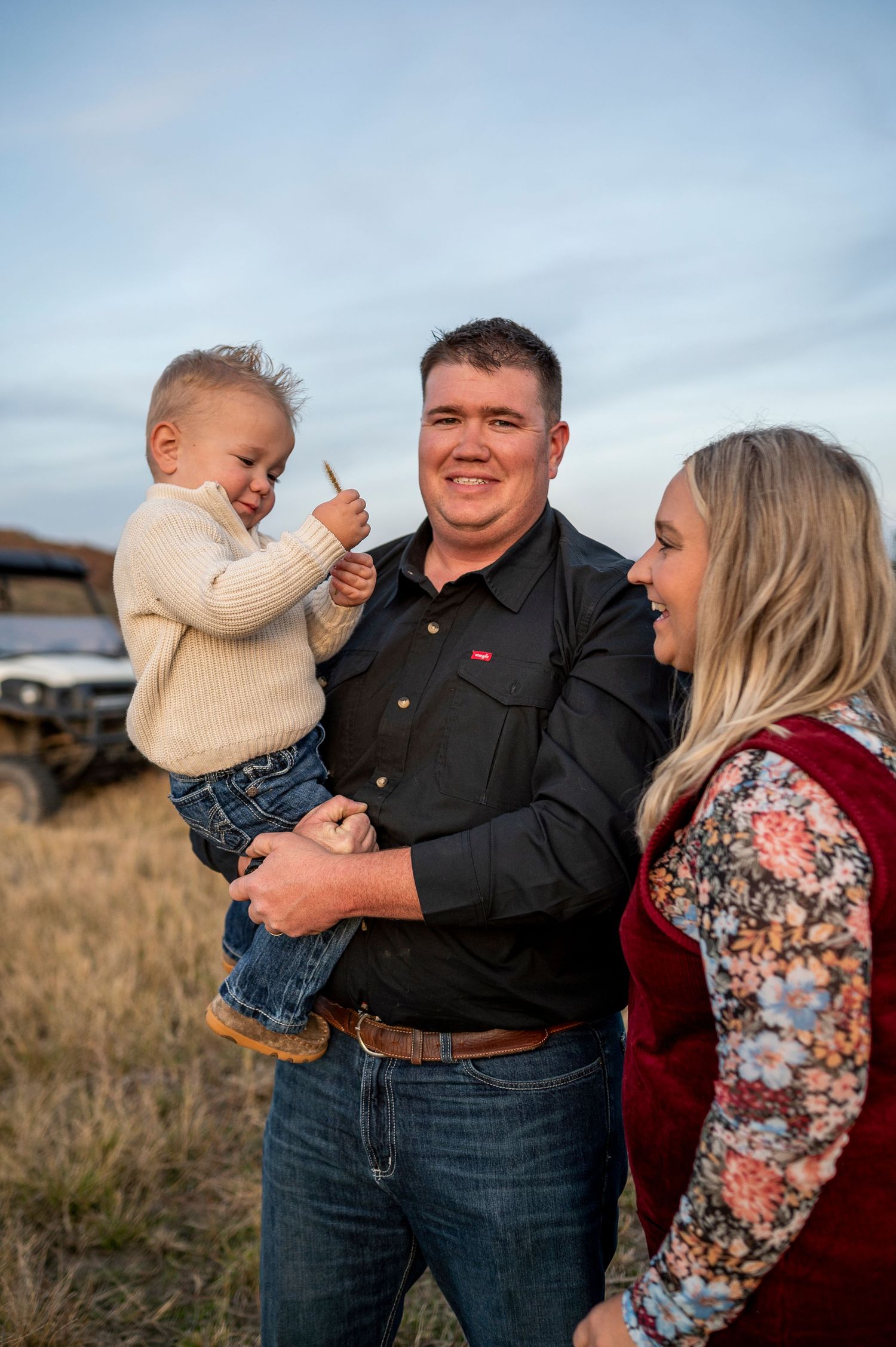 Smiling man in black shirt holds toddler while woman in red vest laughs outdoors near a utility vehicle.