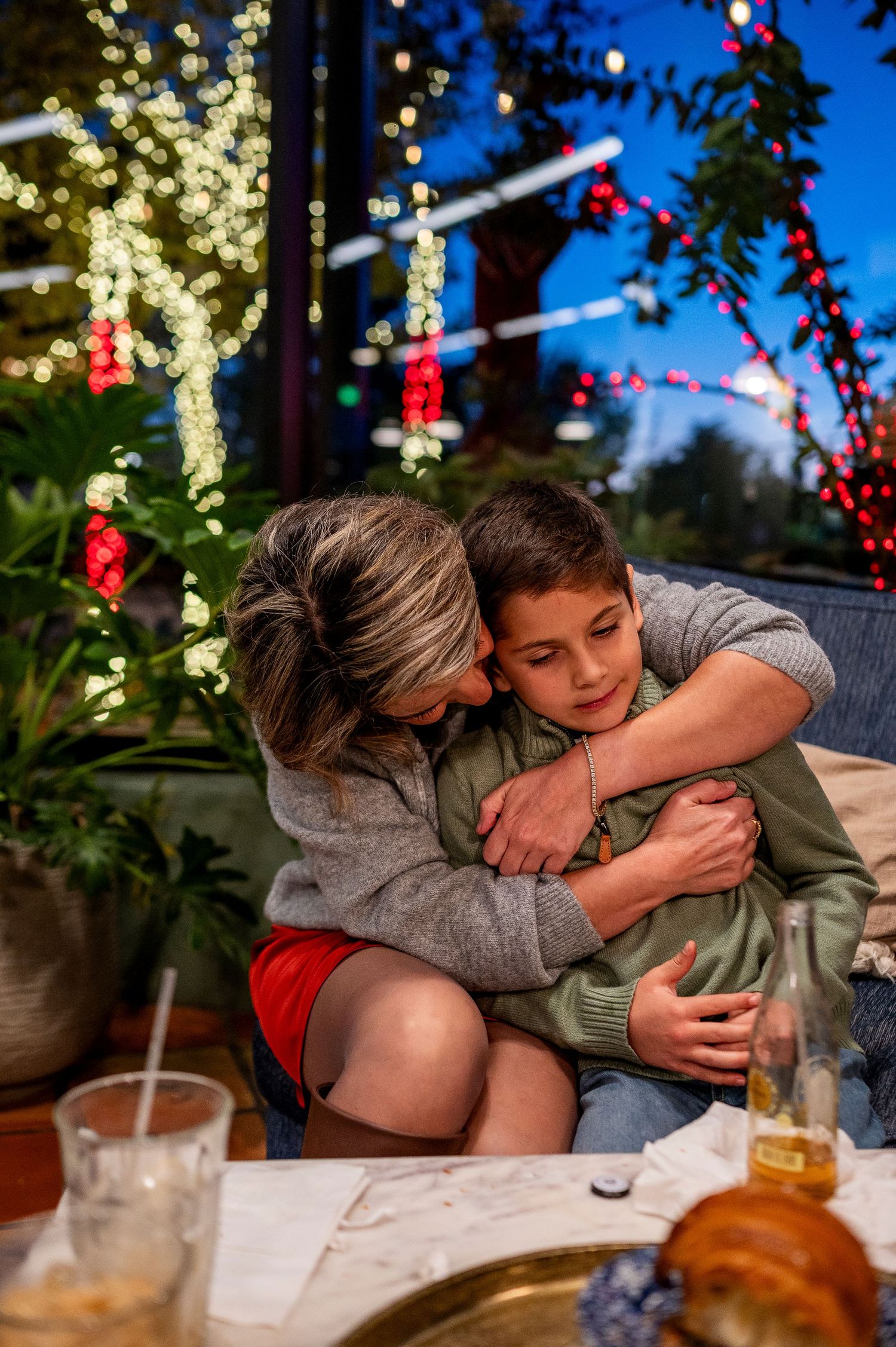A mother hugs her son warmly at an outdoor evening gathering with colorful holiday lights in the background.