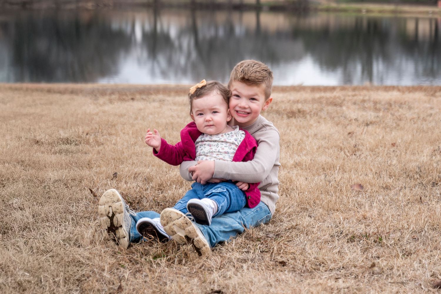 Two children sitting together on brown grass near a lake.