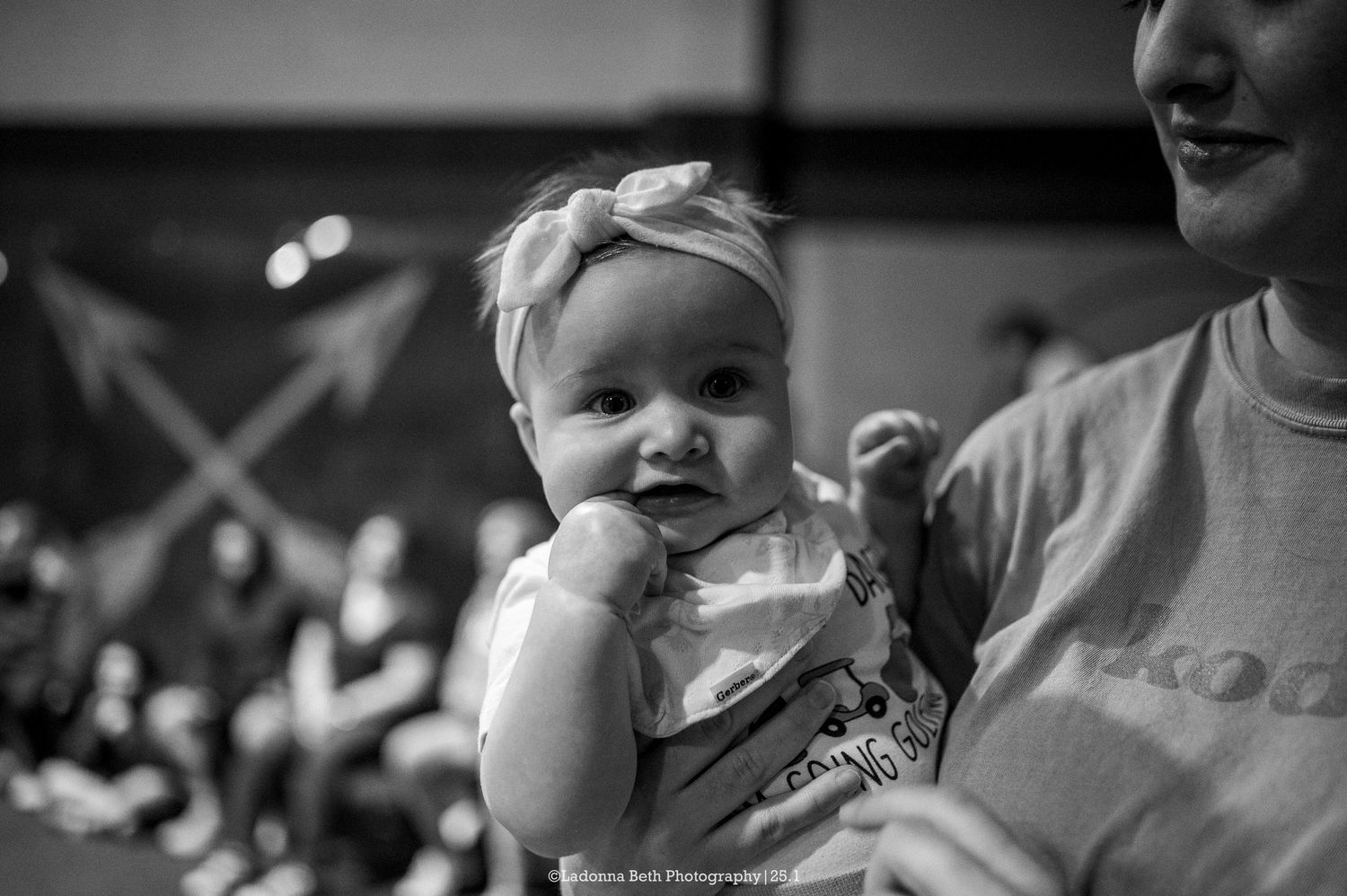 A small baby wearing a bow headband smiles while being held at an indoor event.