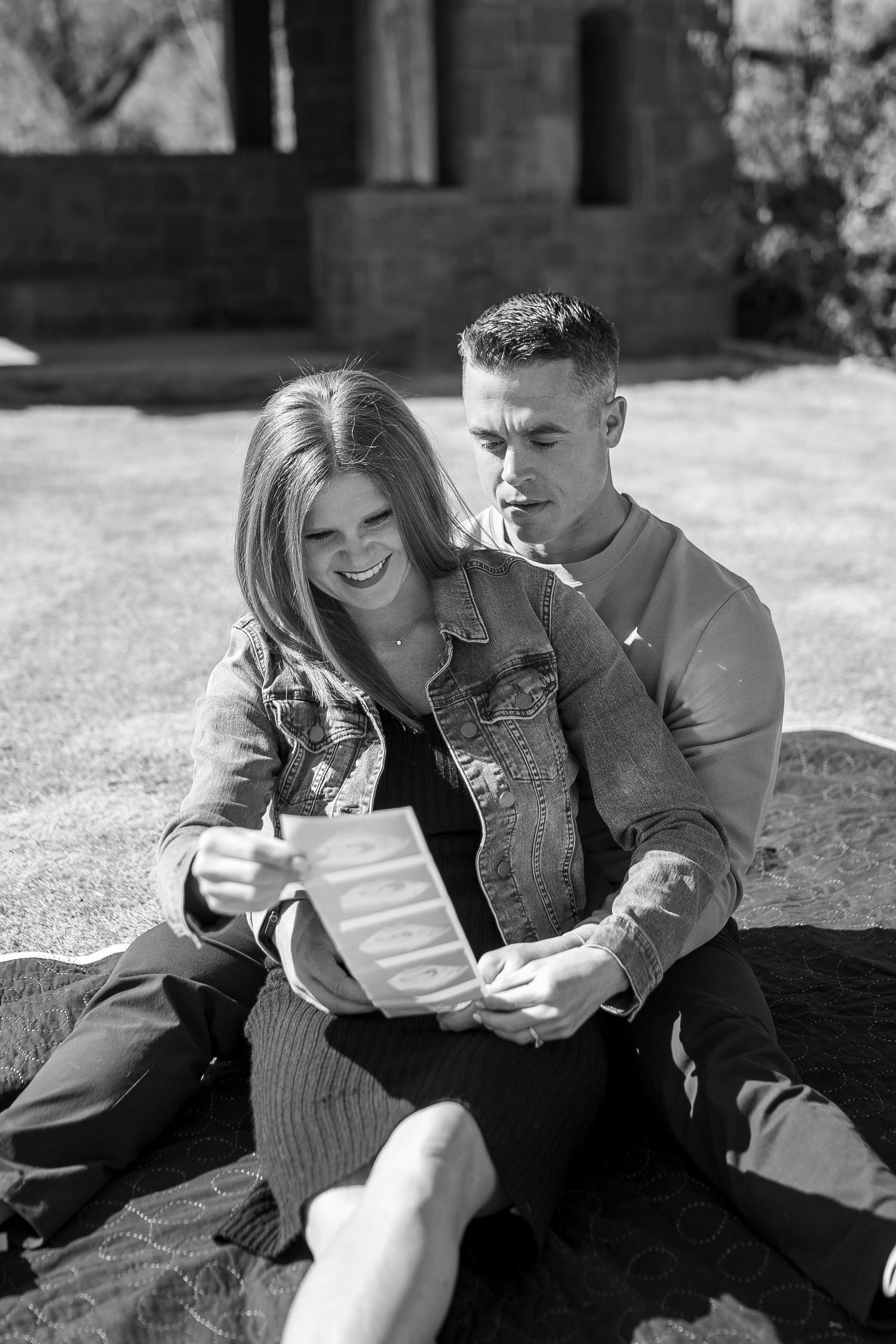 A casual outdoor photo session showing a couple looking at papers while sitting on grass.