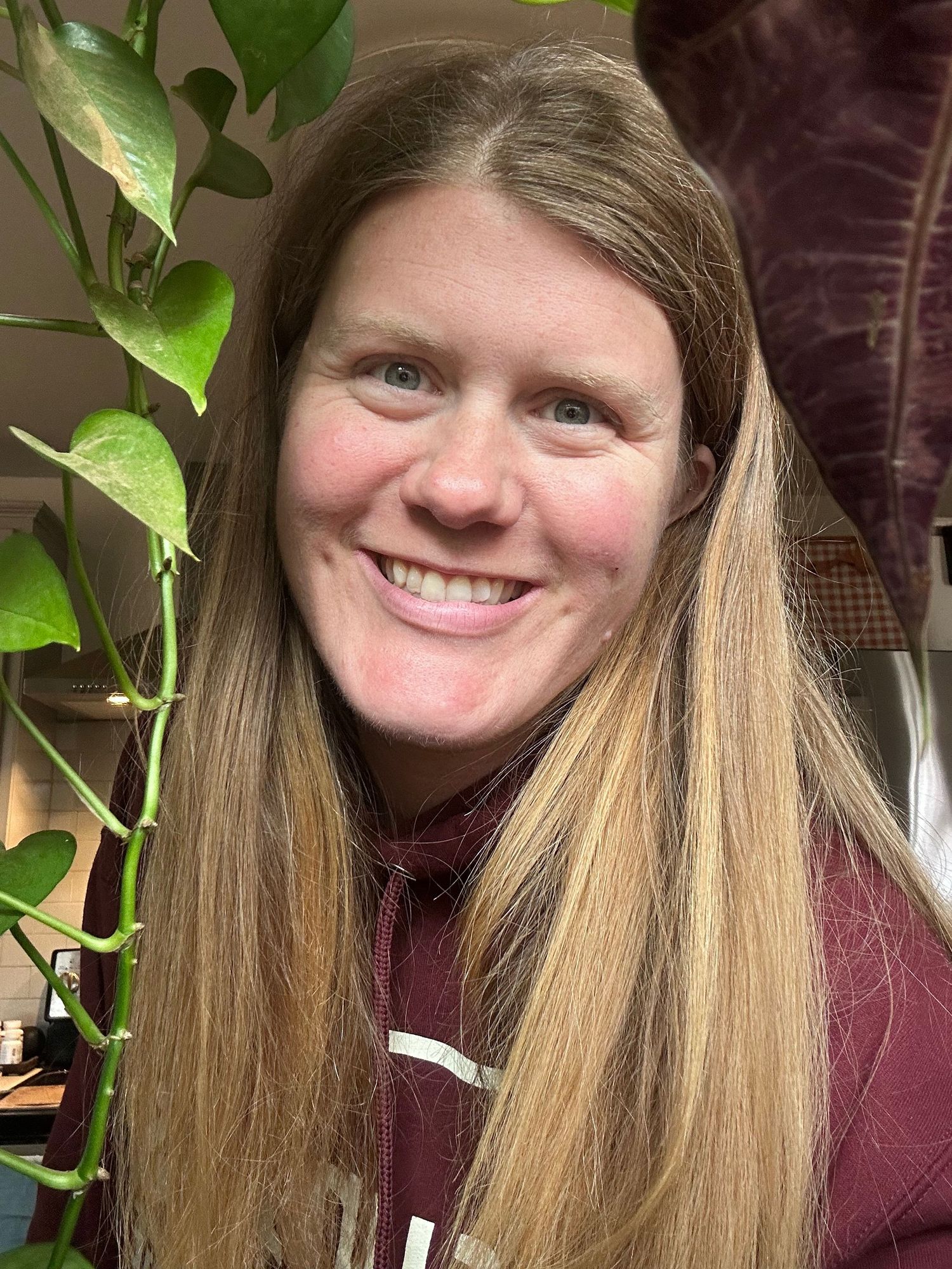 A person smiles warmly next to a potted plant with bright green leaves.