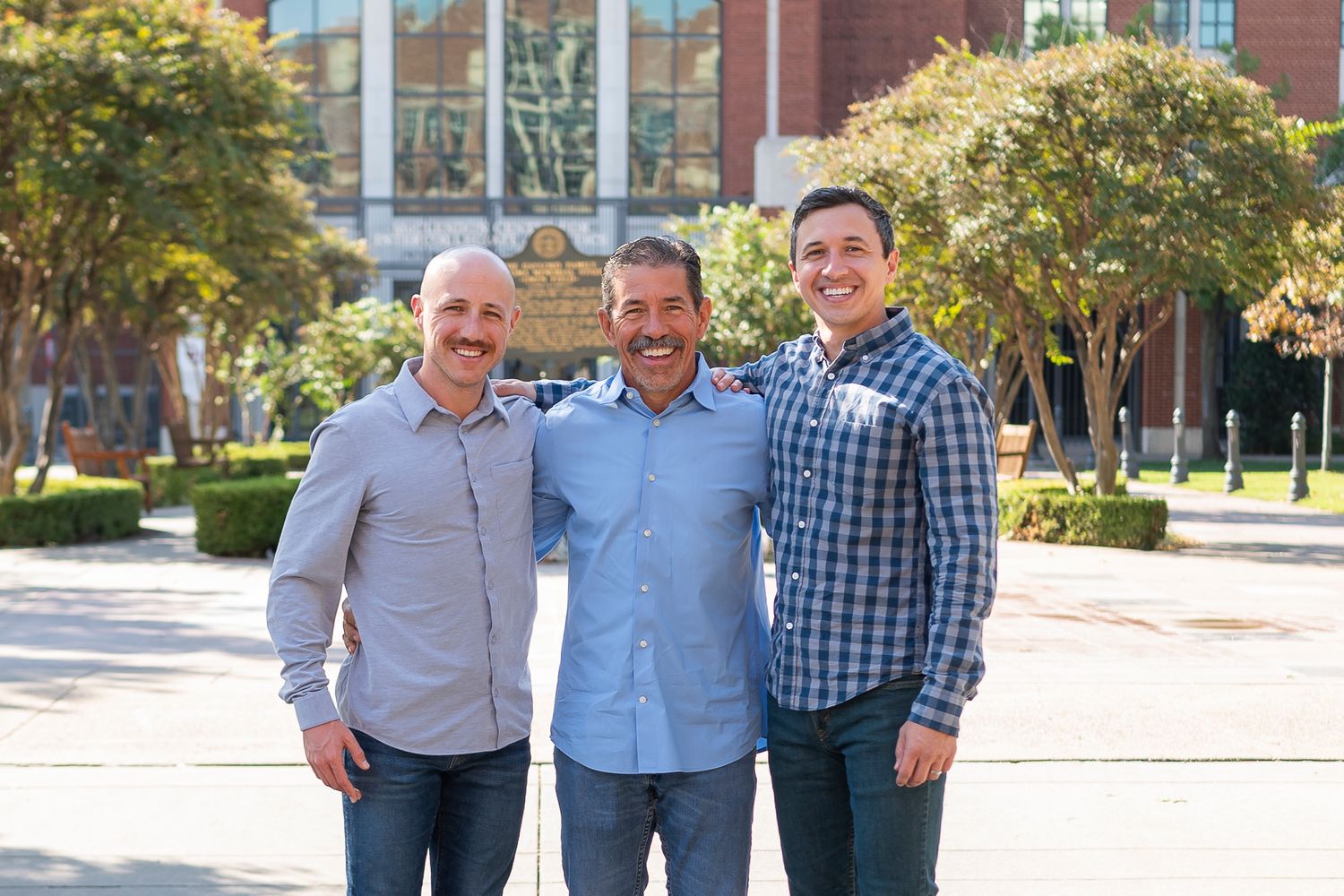 Three men stand together smiling in front of a brick university building.