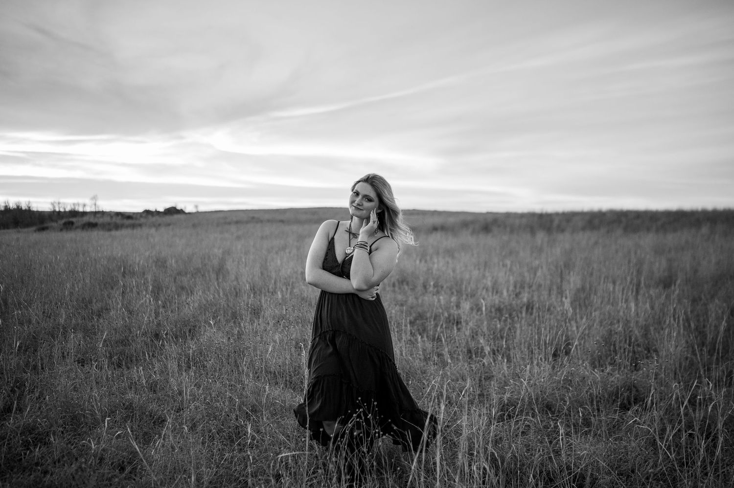Black and white photo of a woman in a long dress standing in a field, smiling softly at dusk.