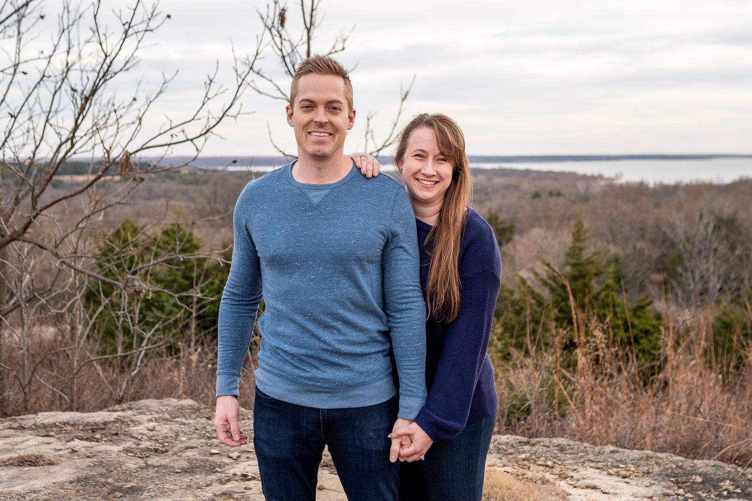 A smiling couple holds hands on a rocky overlook with a scenic winter landscape and water view behind them.