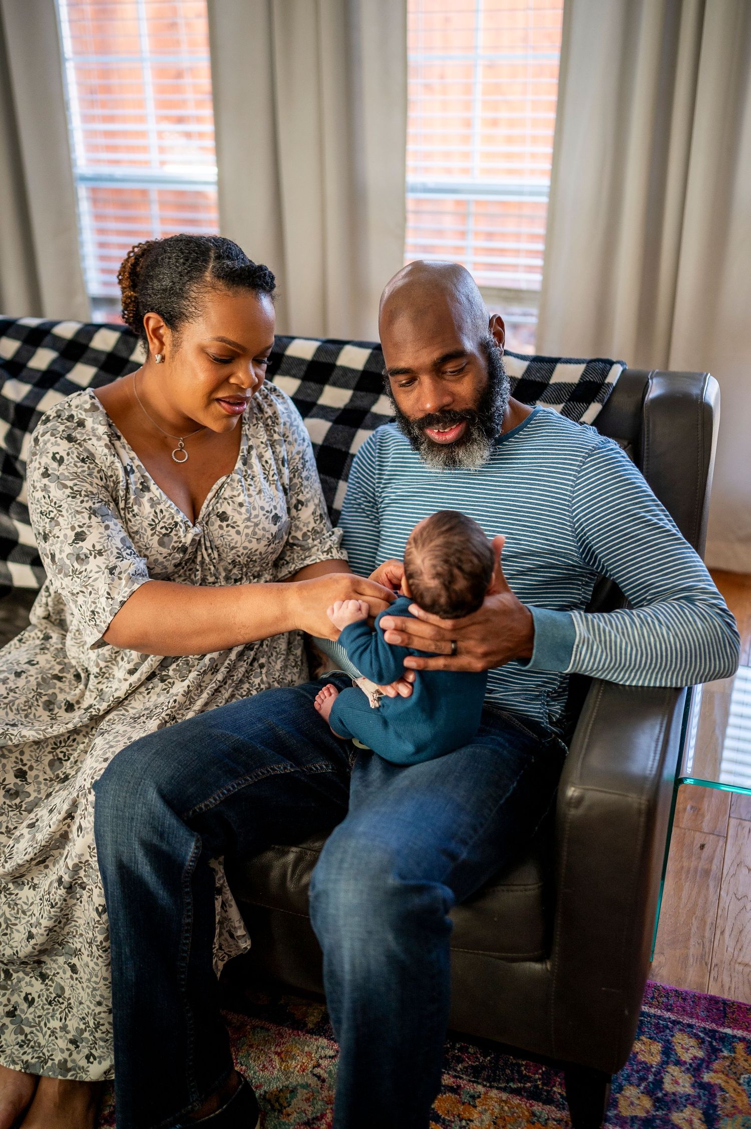 A couple sits together in a cozy living room, lovingly cradling their newborn baby in a leather armchair.