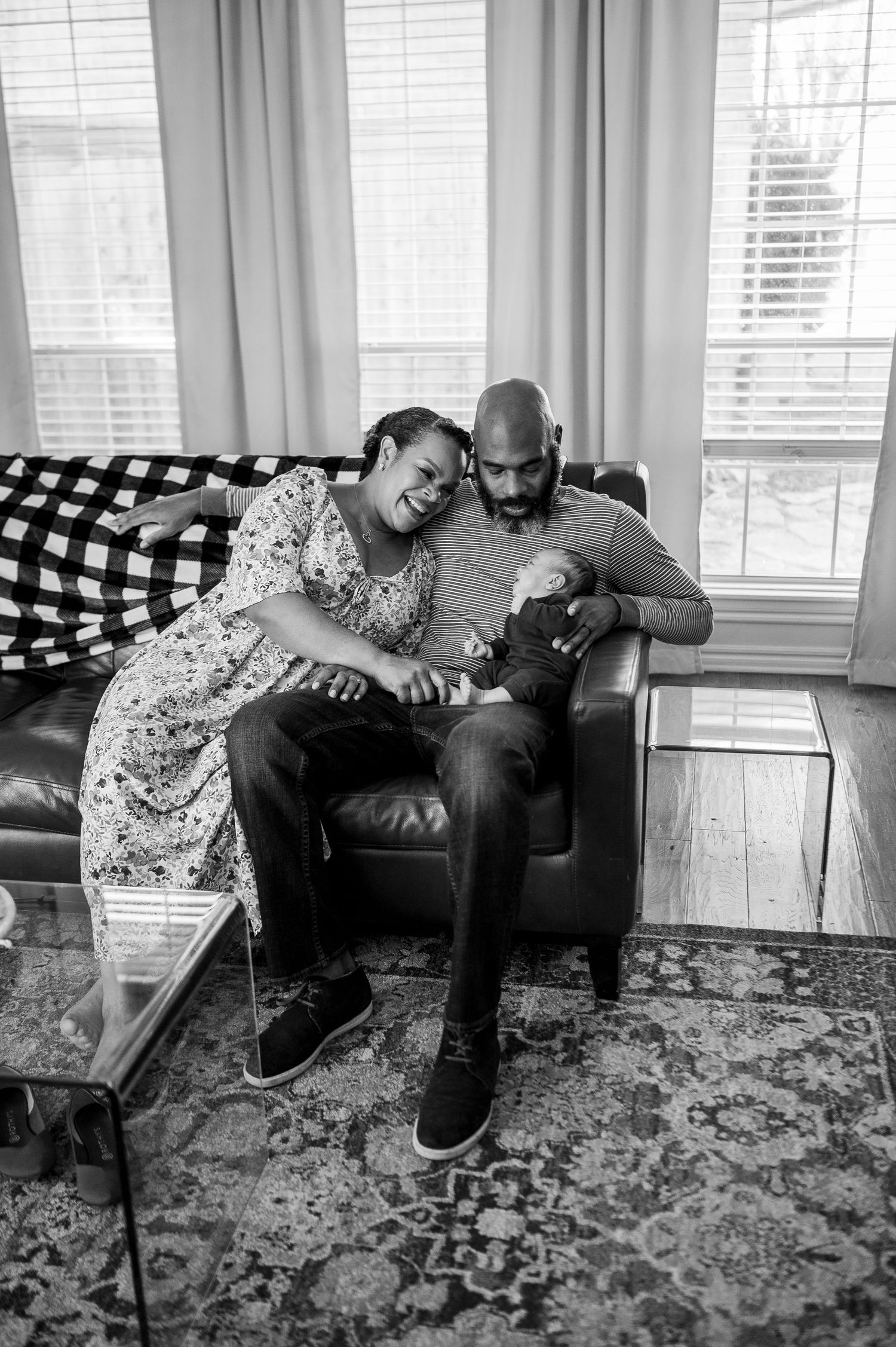 Black and white photo of a couple sitting on a leather chair, tenderly holding their newborn baby in a cozy living room.