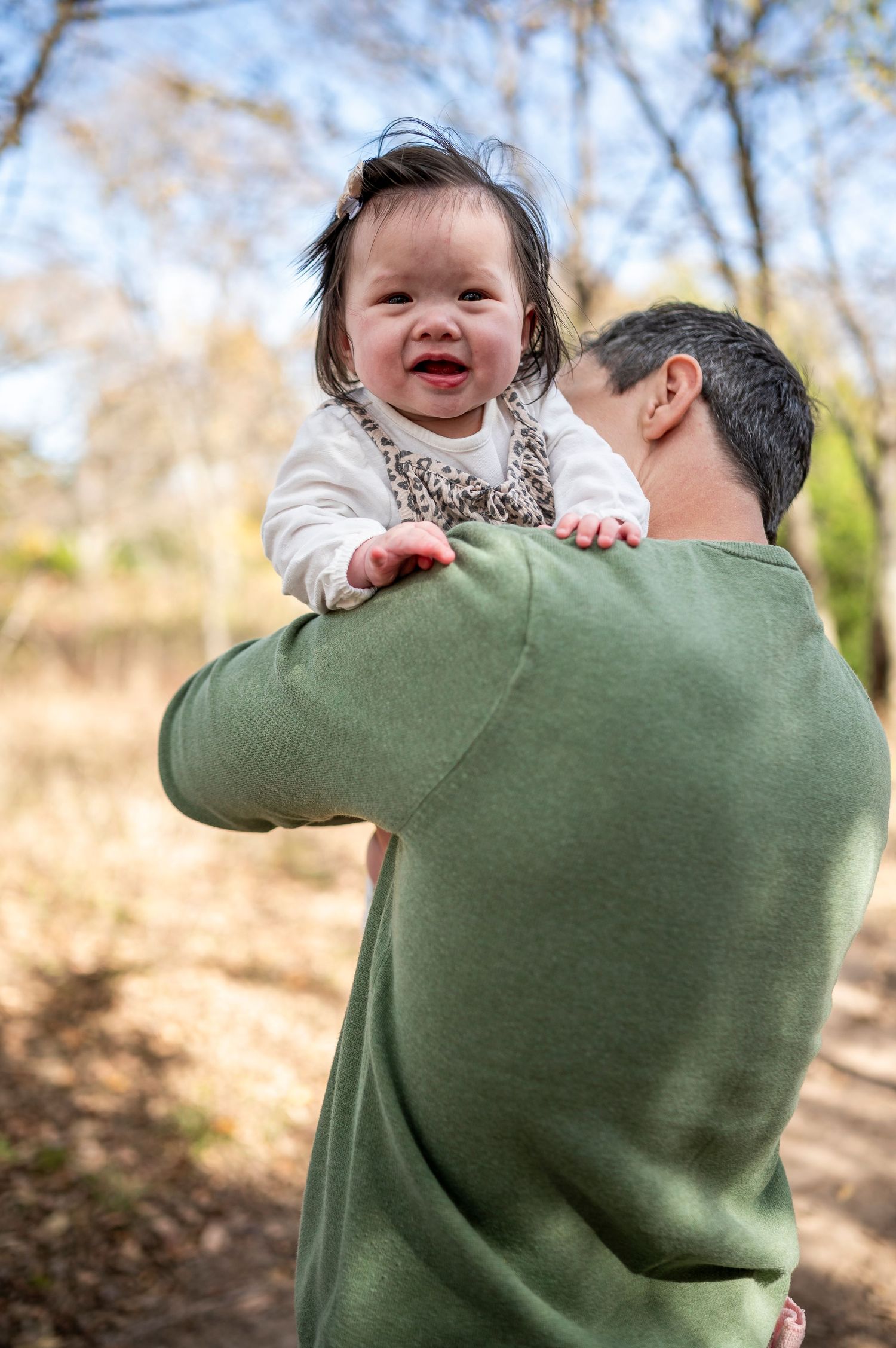 A father in a green sweater holds his baby girl up in an outdoor autumn setting.