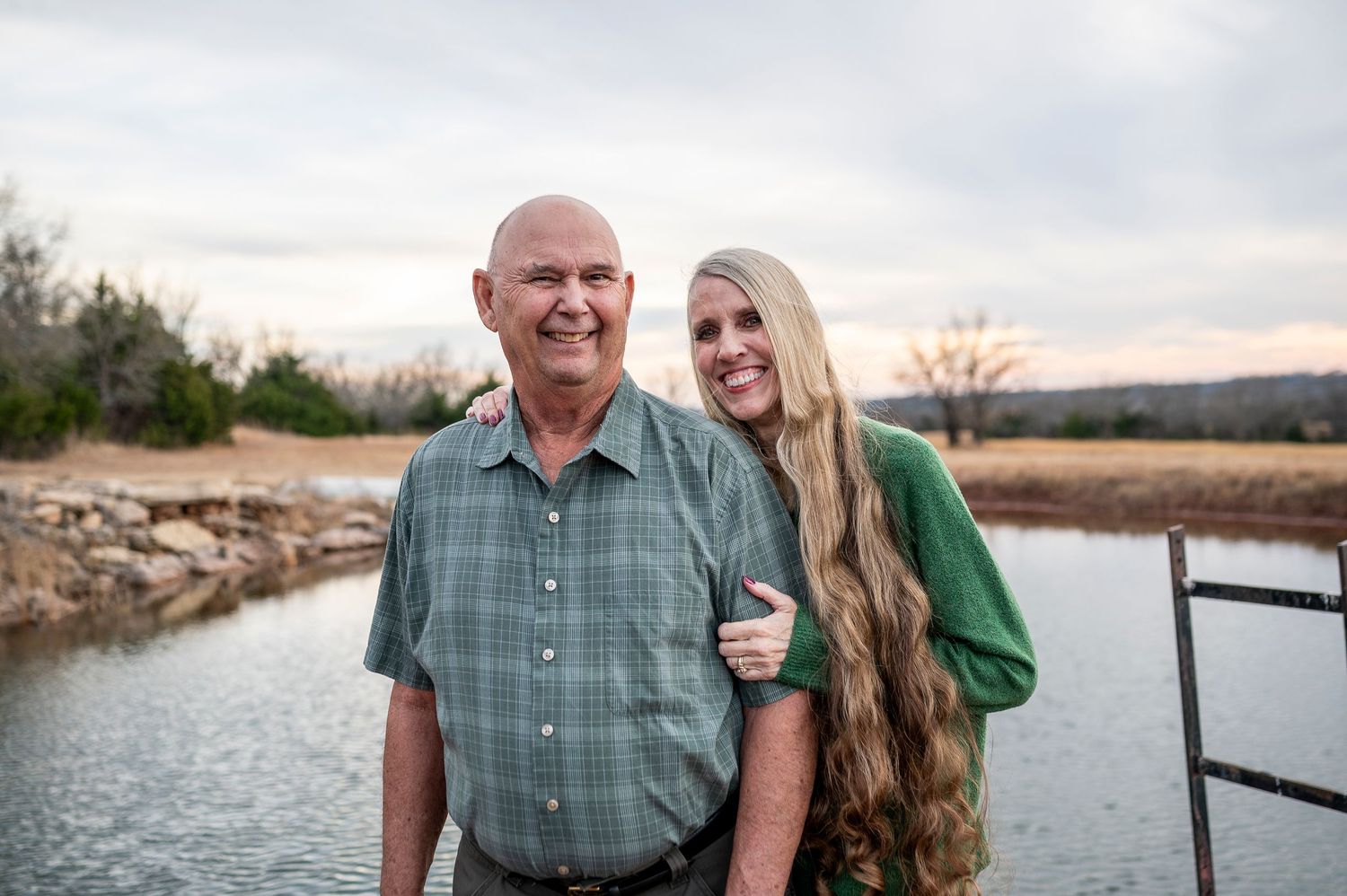An older man and young blonde woman smile together outdoors near a serene river at dusk.