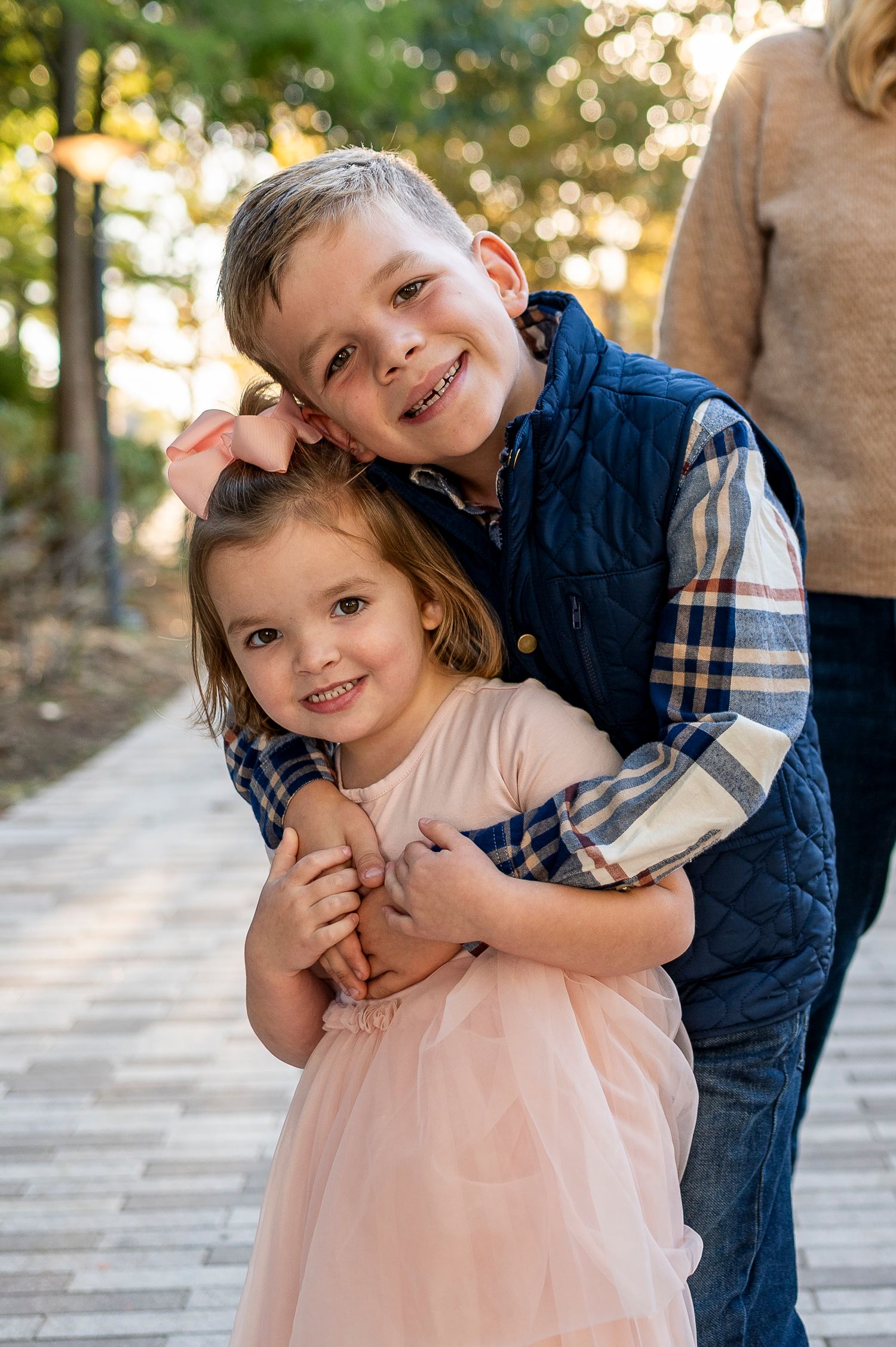 Boy in navy vest hugs smiling little girl in pink tutu dress outdoors on a stone path.