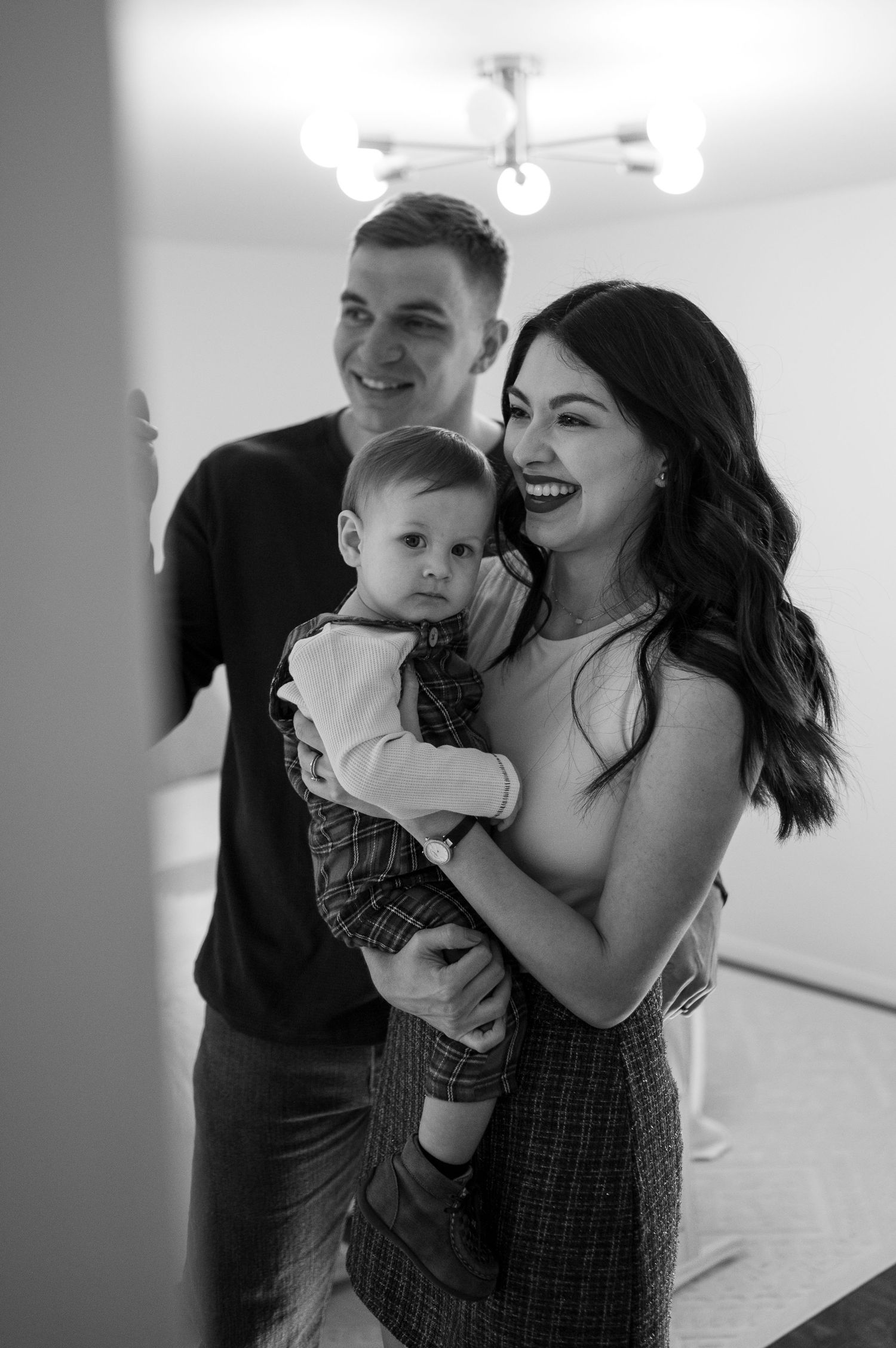 A candid black and white portrait of a happy family moment with their baby in plaid overalls.