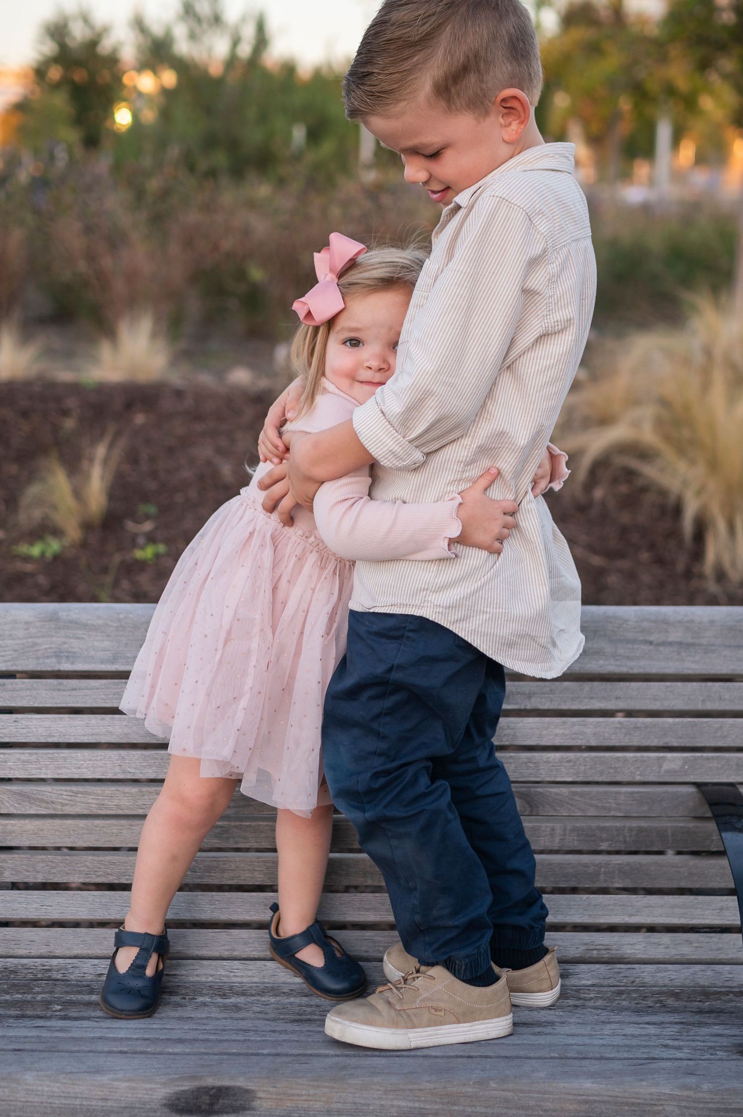 Two children embrace while standing on a wooden deck with ornamental grasses in the background.