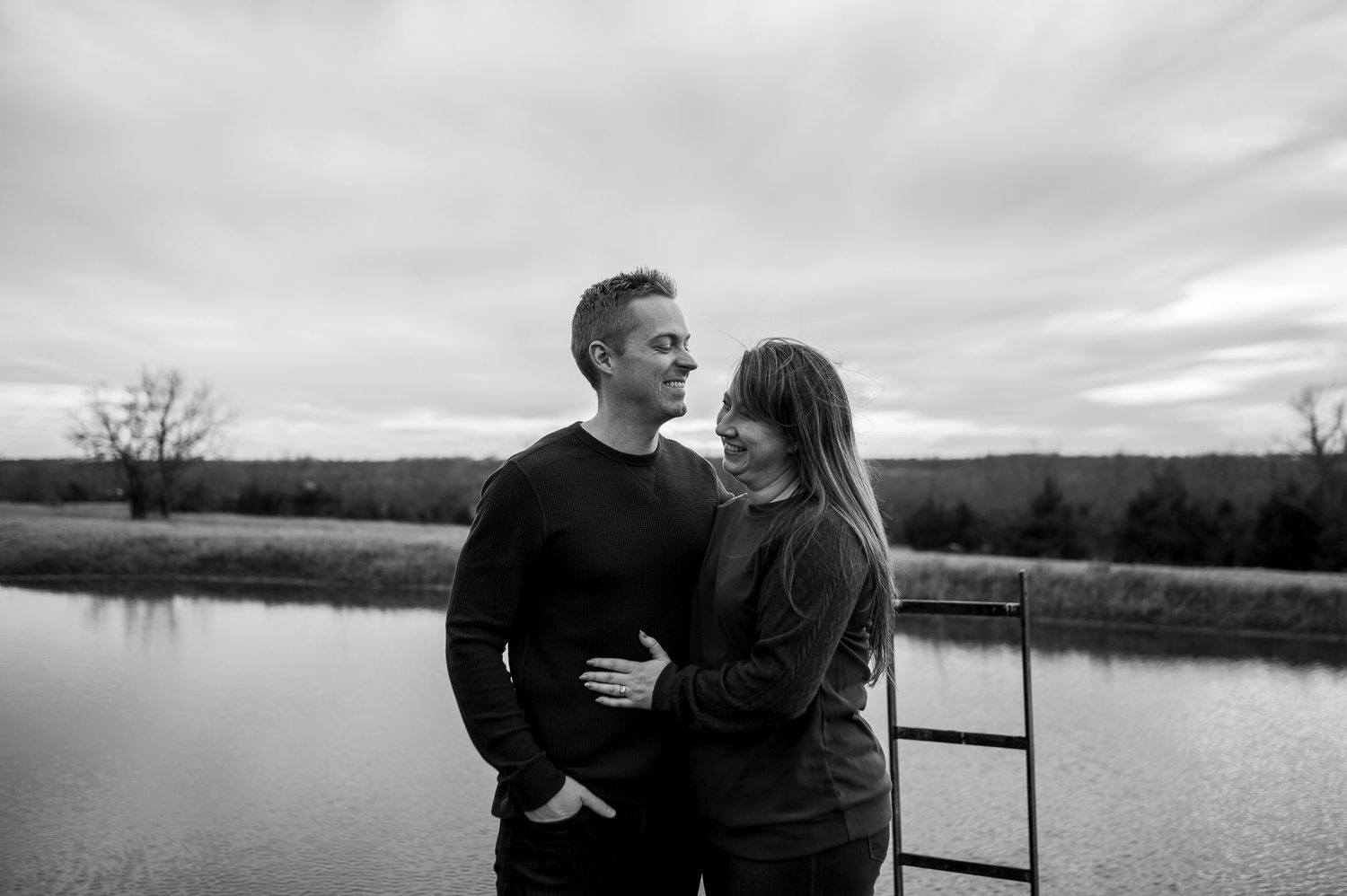 Black and white photo of a couple embracing near a serene pond with a cloudy sky backdrop.
