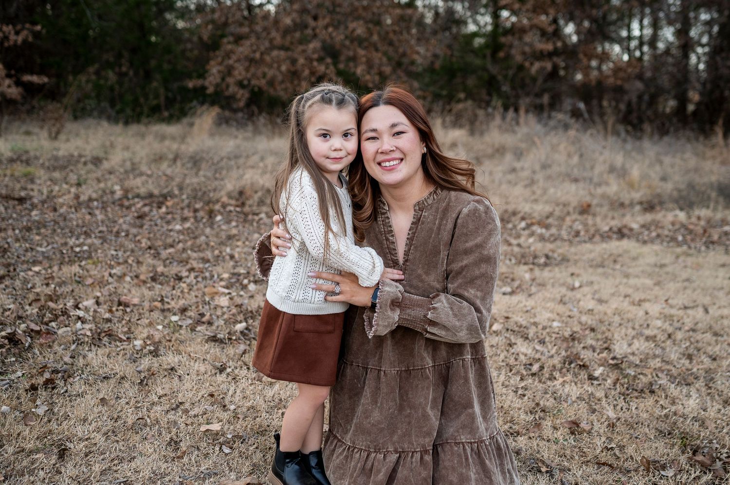 A smiling woman in a brown dress holds a young girl in a white top and skirt outdoors in a fall field.