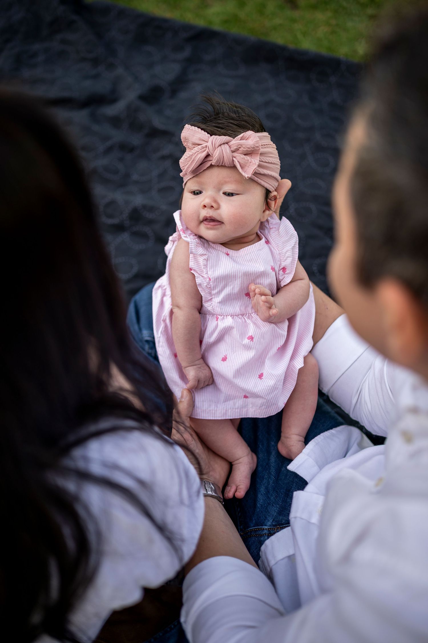 A baby wearing a pink dress with a bow headband lies on a cushioned surface while adult hands support them.
