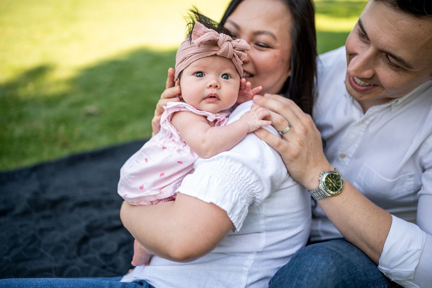 A baby in pink clothing enjoys a tender outdoor moment with parents on a dark blanket in a sunny park.
