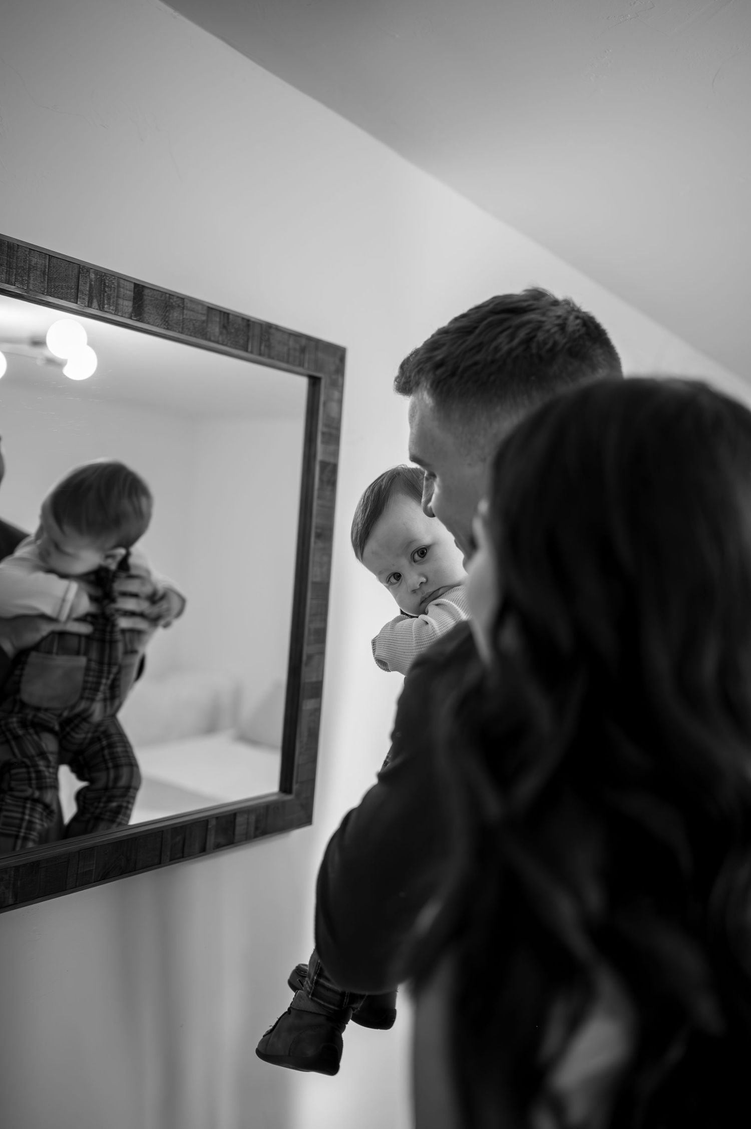 A family looks at their reflection in a mirror while holding their baby in a black and white photograph.