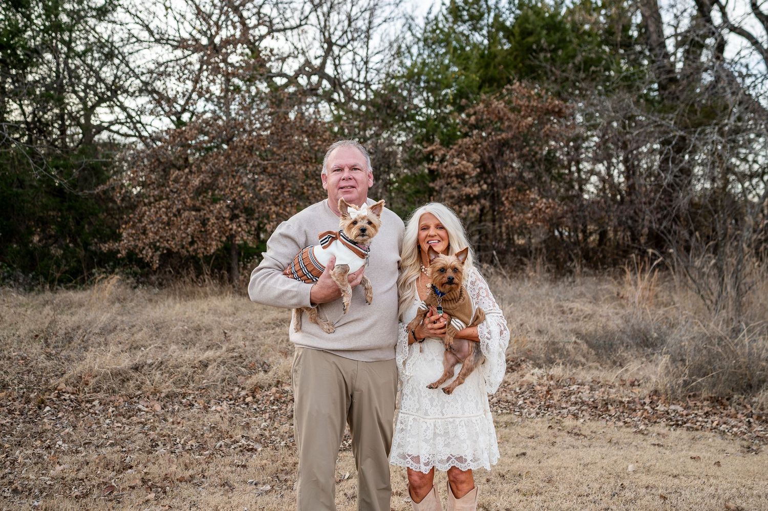 Couple holding small dogs in a rustic outdoor setting with dry grass and bare trees in the background.