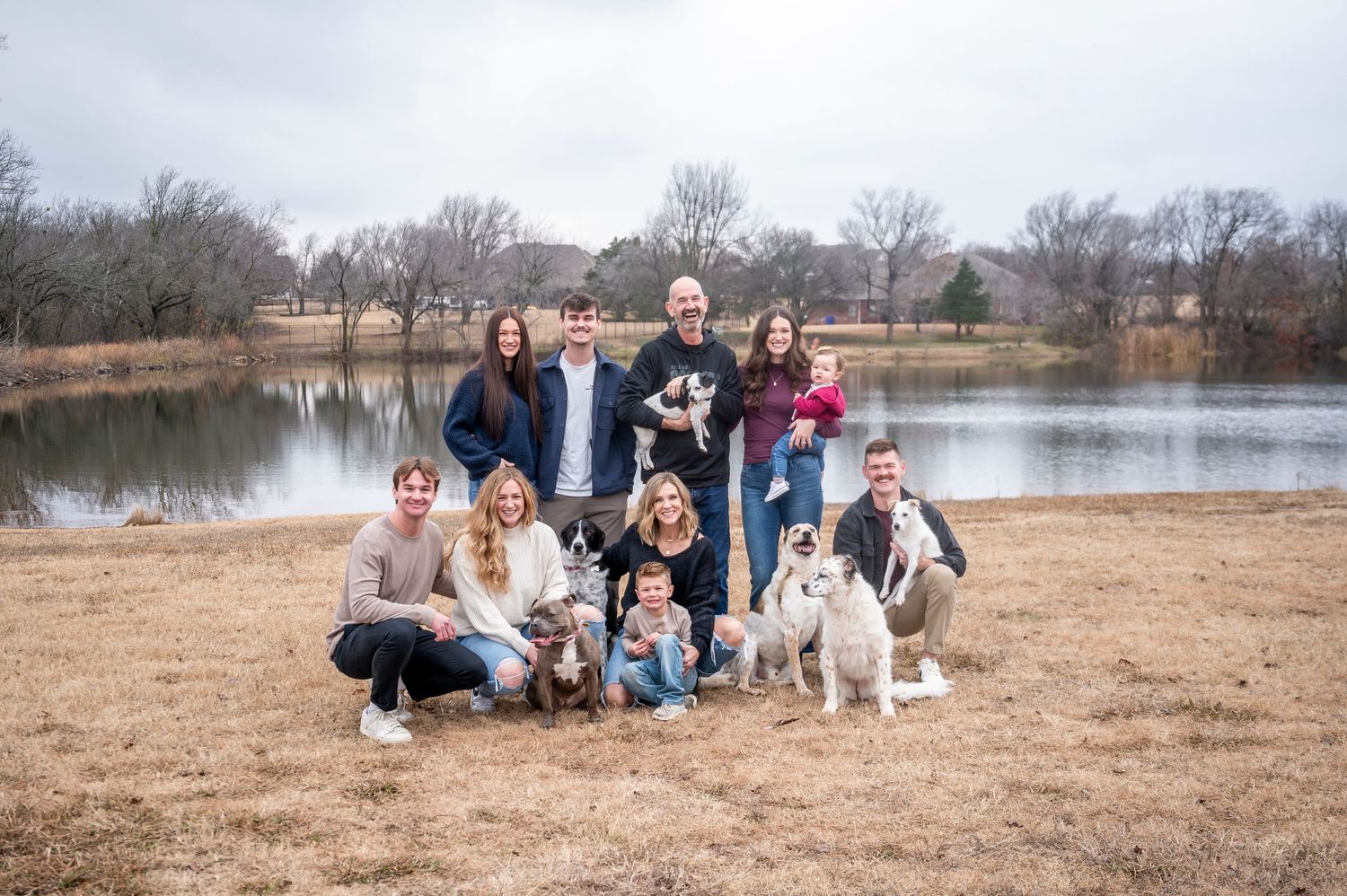 Large group photo with multiple people and dogs gathered by a lake.