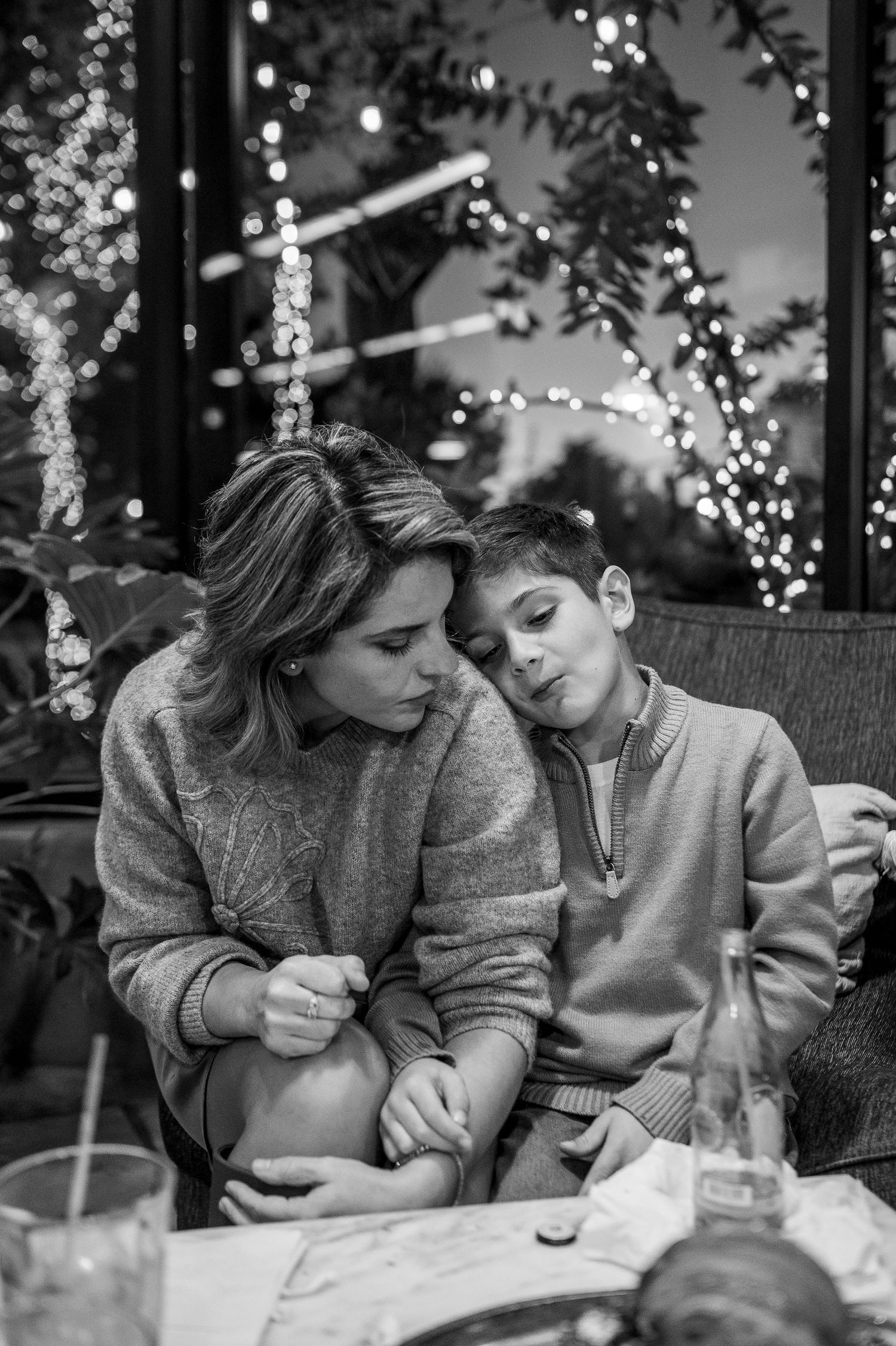 Black and white photo of a mother and son sharing a tender moment at a table with bokeh lights in the background.