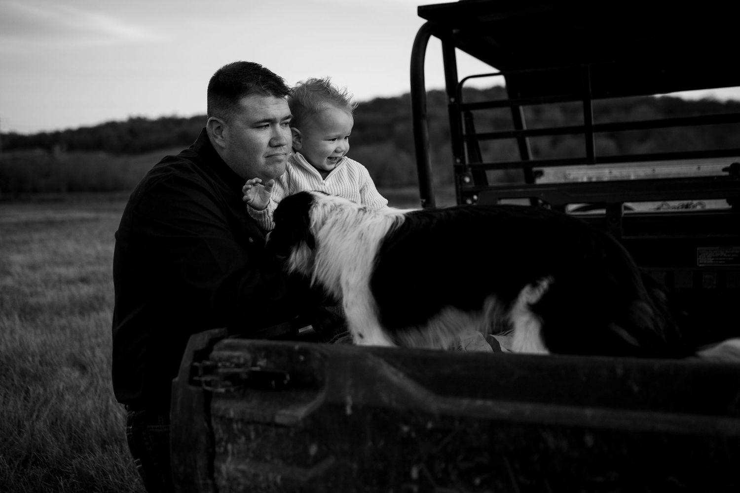 Black and white photo of a man holding a baby near a truck tailgate with a border collie dog in a rural field.