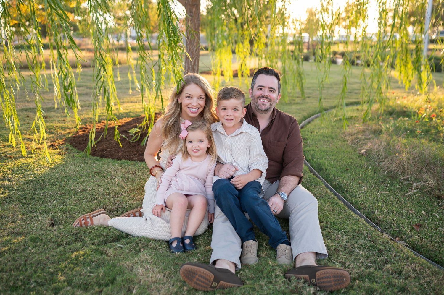 Family sitting together on rustic wooden bench surrounded by willow trees at sunset.