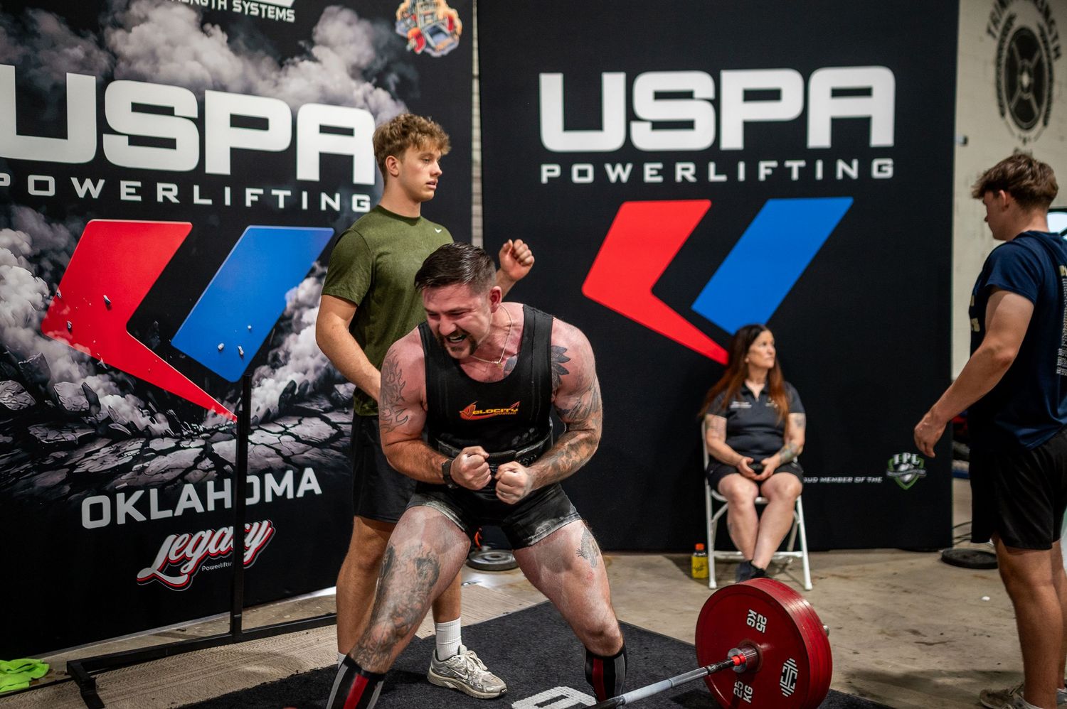 Powerlifter celebrates a lift at a USPA Powerlifting Oklahoma Legacy competition, surrounded by spotters and judges.